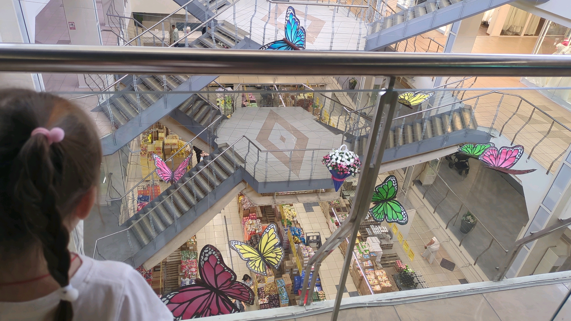 The image depicts the interior of a multi-level commercial establishment, possibly a shopping mall or department store, viewed from an upper level. A young girl with her back to the camera, her hair in a braid, is looking down at the scene. The central focus is a large atrium with multiple staircases and walkways. Decorative elements, in the form of large, colorful paper butterflies, are suspended throughout the atrium. A blue butterfly is prominently positioned near the top of the atrium, while other butterflies in pink, yellow, and green are visible at various levels. A floral arrangement hangs from a walkway. Below, on the ground floor, aisles of merchandise are visible, indicating a retail environment. The architecture features modern construction with metal railings and glass elements. The overall impression is one of a well-lit, open interior space decorated for a festive or seasonal display. Without further information, it is impossible to ascertain the specific location within Chișinău, Moldova.