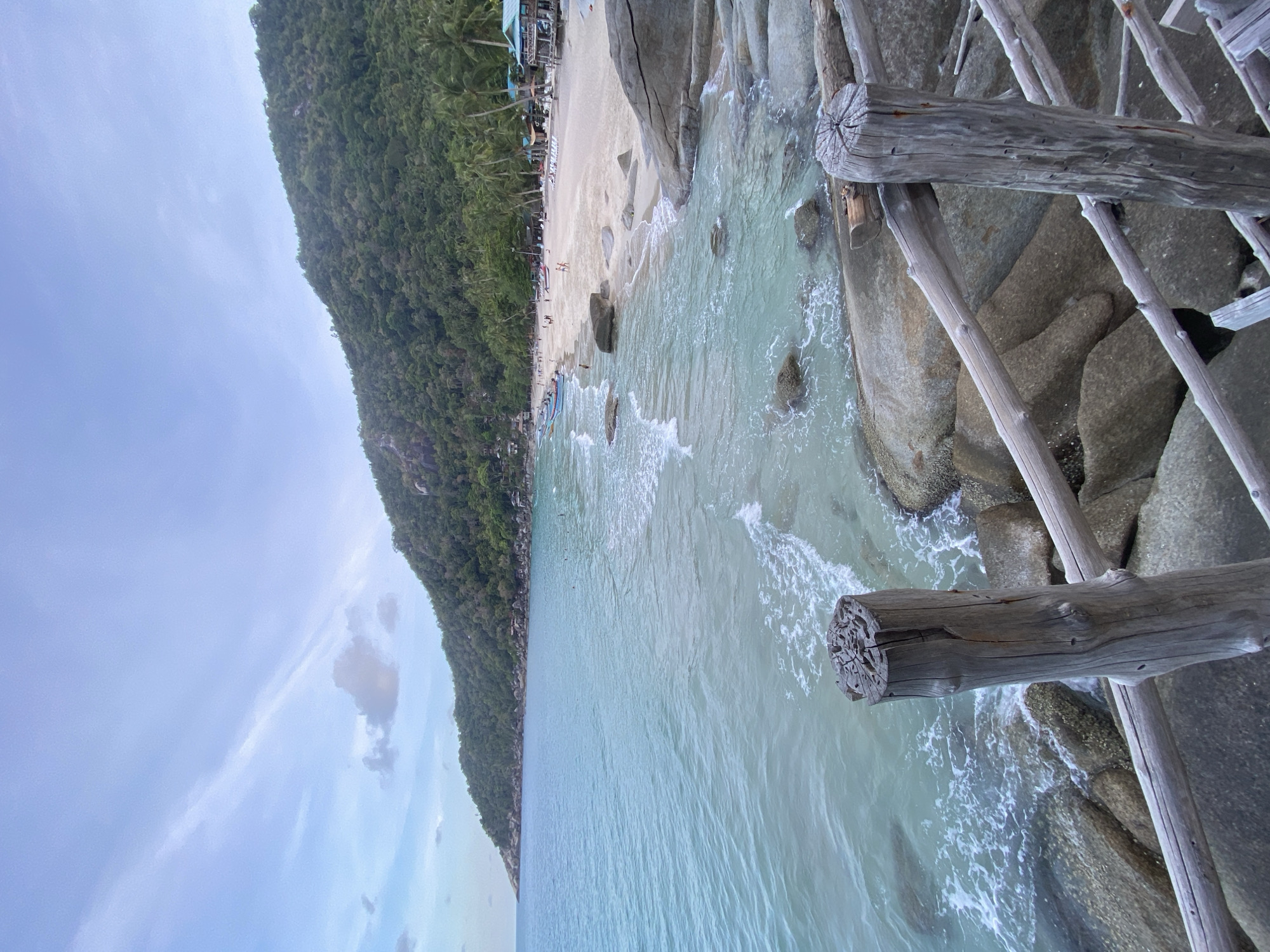 This photograph captures a coastal scene, likely in Ban Haad Rin, Thailand. A beach lined with trees is visible on the left, with a sandy shore populated by people. The water is clear and turquoise, with visible waves breaking against the shore. To the right, a wooden structure, possibly a pier or walkway, extends into the water. The structure is constructed from weathered wooden posts and beams, creating a rustic appearance. Large rocks are visible beneath and around the wooden structure, partially submerged by the water. The sky is overcast with clouds.