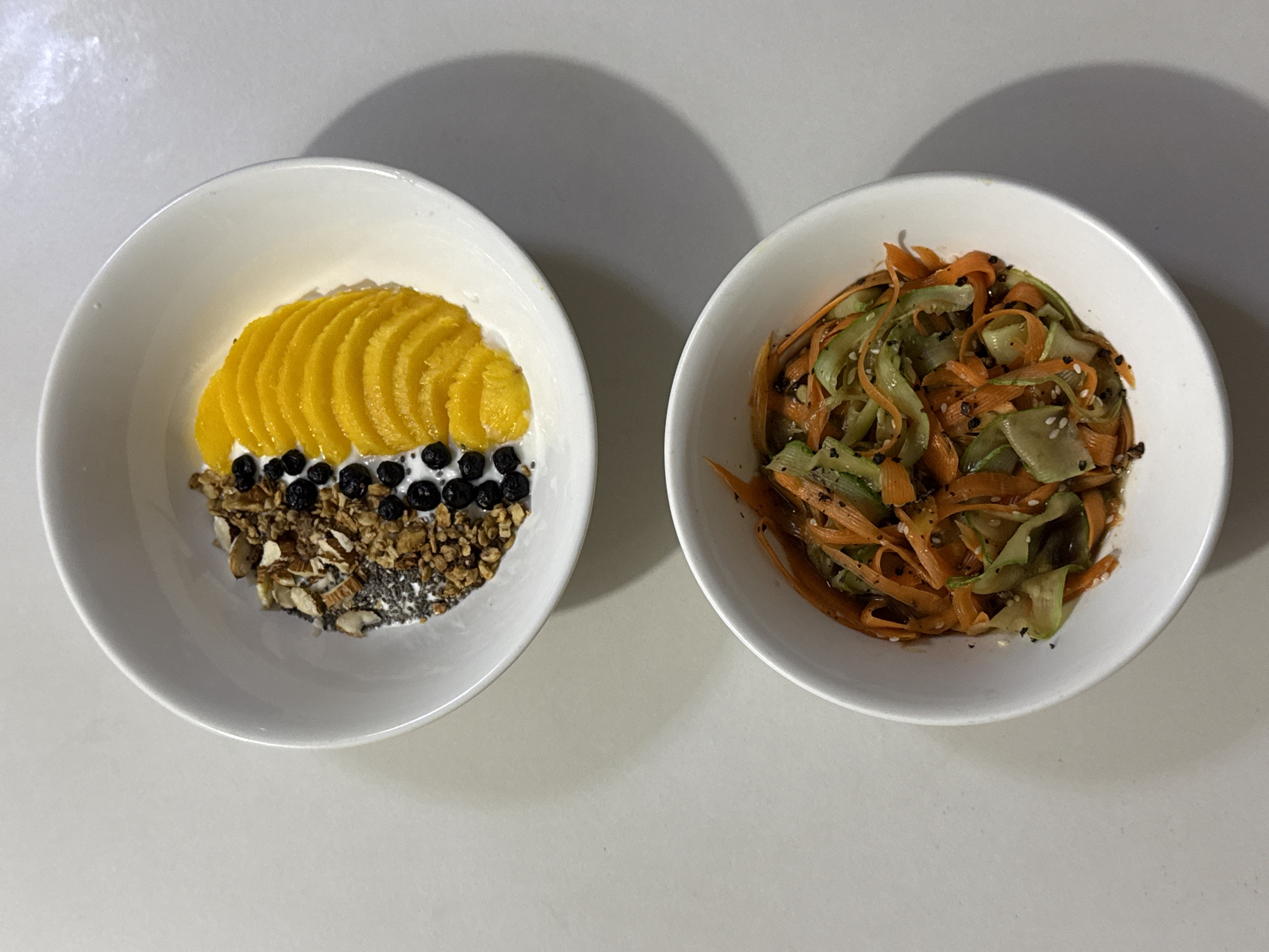 The image displays two white ceramic bowls of food placed on a plain, light-colored surface, with the scene located in Davao City, Philippines. The top bowl contains a yogurt or cream-based dish divided into sections. The left section features a mix of granola, chopped nuts (resembling almonds and walnuts), and small dark chia seeds. Adjacent to this, a vertical line of small, dark blueberries is arranged. The right section of the top bowl contains bright yellow, thinly sliced mango pieces, uniformly arranged in an overlapping fan pattern. The bottom bowl holds a salad-like preparation composed of ribbon-cut vegetables. This includes bright orange strips, likely carrots, and pale green strips, likely cucumbers, all coated in a light, glossy dressing. White sesame seeds and smaller, dark flakes or seeds are sprinkled over the vegetable dish. No people are visible in the scene.
