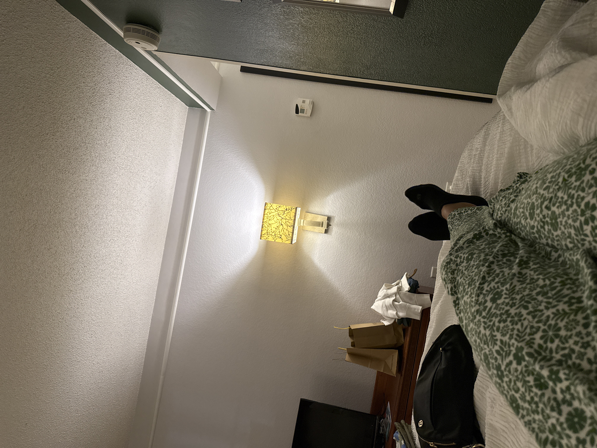 The photograph appears to be taken inside a hotel room, likely in San Antonio, United States of America, based on the prompt. The angle is looking upwards from the bed.
The visible components are:
* **Wall and Ceiling:** A light-colored wall and ceiling with a textured surface.
* **Lighting:** A wall-mounted lamp is present.
* **Thermostat:** A digital thermostat is visible on the wall.
* **Items on a Table:** There are paper bags, some white clothing, and other unidentified items on a wooden table next to the bed.
* **Television:** A television is positioned against the wall near the table.
* **Bed:** A bed with white bedding is visible, extending into the foreground of the shot.
* **Person:** A person is in the bed, wearing socks and a green patterned garment.
* **Bag:** A black bag is located on the bed.
