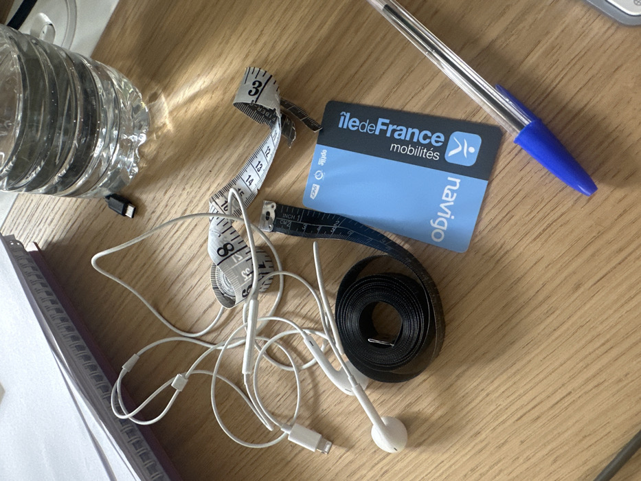 The image displays a collection of personal items resting on a light brown wooden surface. In the upper left, a stack of white papers or notebooks with a visible spiral binding is partially present. A tangled assembly of white wired earphones, featuring a Lightning connector, occupies the left-central area. To the right of the earphones, a white fabric tape measure is partially uncoiled, displaying black numerical markings. Below this, a black fabric tape measure is tightly coiled with a small silver pin or clip inserted into its center. Adjacent to the black tape measure, a short, blue and black measuring tape with "INCH" markings is visible. To the immediate right of the measuring tapes lies a light blue Navigo card, bearing the text "Navigo" and "îledeFrance mobilités" along with a stylized human figure logo, identifying it as a public transport pass for the Île-de-France region, specifically Paris, France. In the upper right corner, a clear plastic water bottle is partially visible, containing water, positioned near a small black USB cable with a metallic connector. A translucent blue ballpoint pen rests diagonally across the lower right portion of the frame. The items are casually dispersed across the wooden surface.