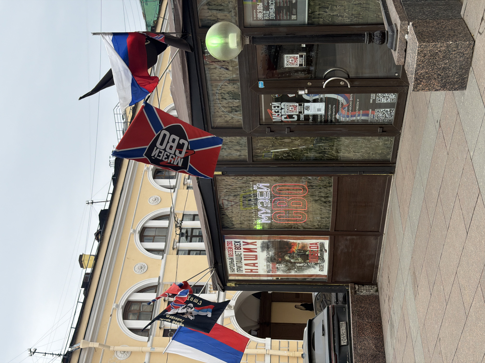 This image depicts a street scene in Saint Petersburg, Russia. The central focus is the entrance to a "Музей СВО" (Museum SVO), referring to the "Special Military Operation." The storefront features dark brown frames and large glass windows. A red neon sign inside spells "МУЗЕЙ СВО." On the left window, a large poster displays soldiers in combat with Cyrillic text translating to "PEOPLE'S MUSEUM SVO INVITES ALL OURS" and "VICTORY WILL BE OURS, BROTHER!" The entrance door includes several smaller informational posters with QR codes.
Above the entrance, four flags are displayed: the national flag of Russia (white, blue, red horizontal stripes), a red flag with a blue diagonal cross and "МУЗЕЙ СВО" text, a black flag featuring a white wolf head, and a plain black flag.
In the background, a light-yellow two-story building with white architectural trim presents multiple arched windows with dark frames. The ground is paved with rectangular tiles in varying shades of brown and gray. A dark granite curb and base are visible in the foreground. The rear of a dark gray Geely SUV, bearing Russian license plate Y049CT 198, is partially visible on the left. A street lamp with a spherical, frosted green light fixture stands to the right of the entrance. One individual, appearing to be a female with long blonde hair and dark clothing, is partially visible within a shadowed archway in the background. The sky is uniformly overcast.