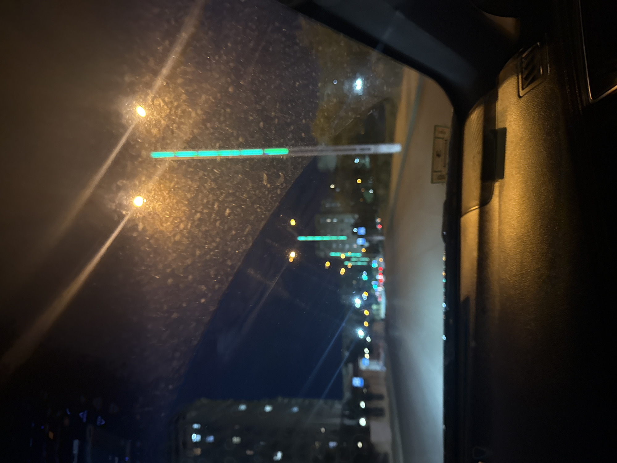 A wide-angle, first-person perspective from inside a vehicle captures a nighttime city street scene. The foreground is dominated by the dark dashboard of the car, reflecting orange and yellow light from streetlights. The windshield is dusty and streaked, with raindrops visible in the upper left corner. Beyond the windshield, a dark blue night sky transitions into a street illuminated by various lights.
On the left side of the street, a tall, modern building with many illuminated windows rises into the dark sky. Further down the street, rows of streetlights line the road, some emitting a warm yellow glow, while others display a vibrant green light from vertical LED elements. Several cars are visible on the road, their headlights and taillights creating blurry streaks of light. The street itself appears to be paved and stretches into the distance. The overall impression is that of a car driving through a city at night, possibly on a rainy evening. The location is identified as Unknown, Unknown.