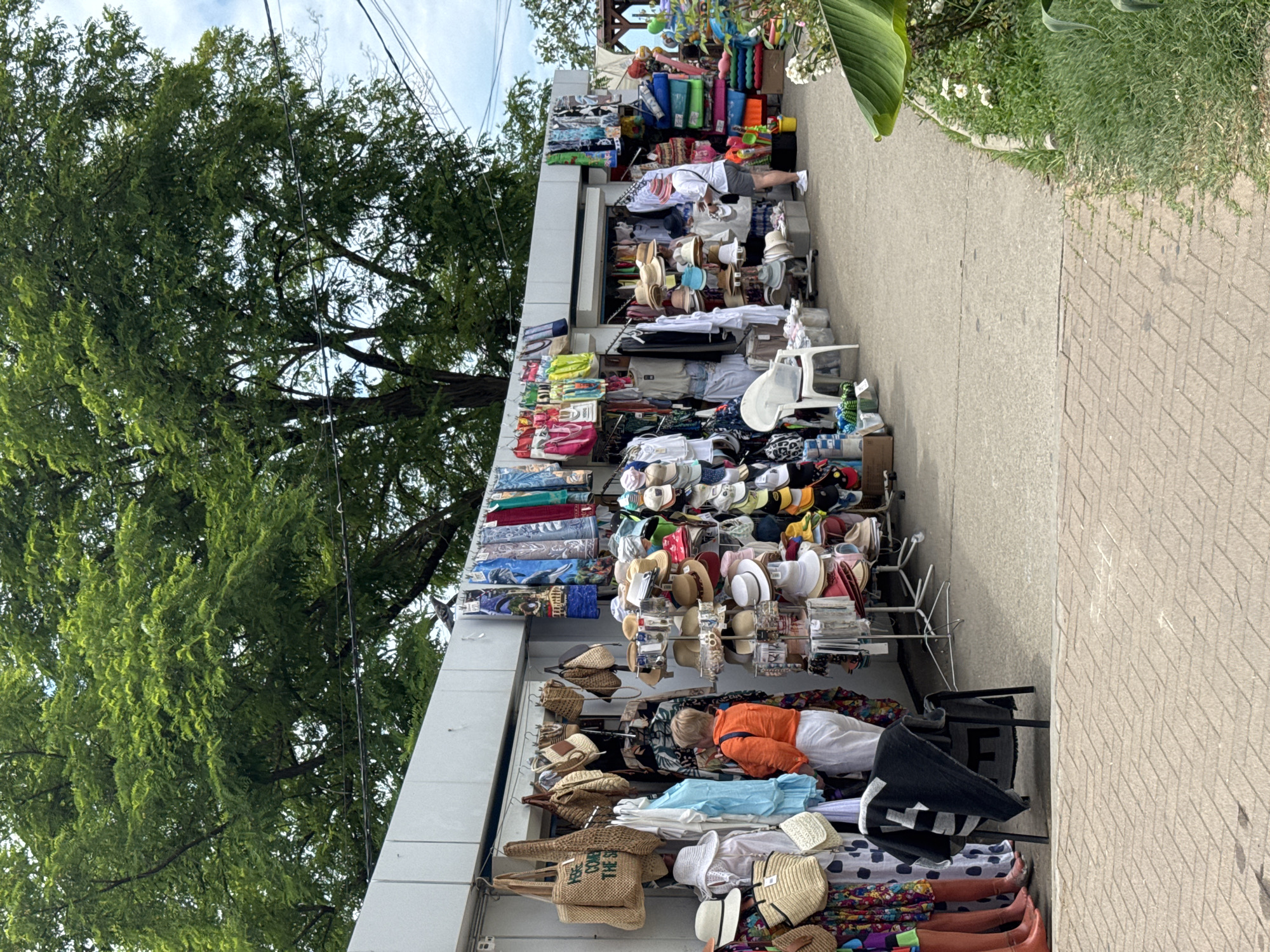 The image depicts an outdoor market stall selling beachwear and accessories, likely in Sochi, Russia, given the style of the market and the warm-weather clothing. In the foreground, a section of a brick pathway is visible. The market stall itself is set up under a white awning. To the left, mannequins display colorful dresses, and several straw bags and white hats are hung. A woman in an orange jacket and white pants is browsing items. Further down the stall, a variety of hats, including straw hats and baseball caps, are displayed on racks. Other clothing items, such as shirts and dresses, are also for sale. In the background, a large green tree provides shade, and electrical wires are visible against a cloudy sky. To the right of the market, a patch of green grass and flowering plants suggest a seaside or park setting. Another person, wearing a white shirt and grey shorts, is looking at items on a rack. Stacked colorful towels and bags are also visible. The overall scene suggests a vibrant tourist area during the day.