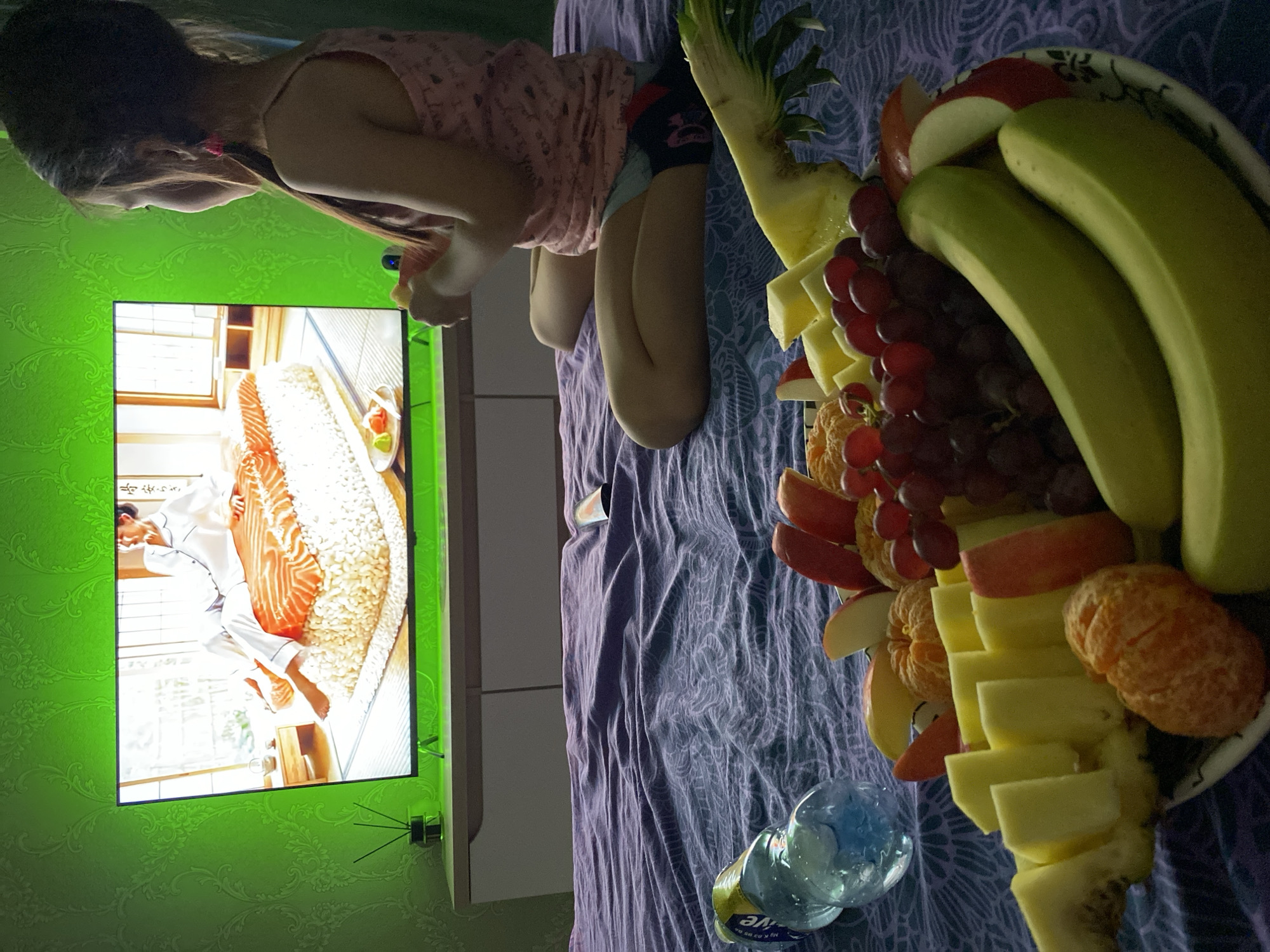 A young girl sits on a bed watching television while a platter of fruit sits beside her in an interior setting, most likely a home.
**People:** One young girl, presumably a child, is visible. She is sitting on a bed, facing a television screen. A man is present on the television screen, and he is wearing a white uniform.
**Objects:** A television is visible on the left. It is attached to a wall that has a patterned green wallpaper. The television is displaying content. There is a large platter of fruit on the bed to the right of the girl. The fruit appears to include bananas, apples, grapes, pineapple, and oranges. A bottle of water is also on the bed.
**Actions:** The girl appears to be watching the television. The man on the television is slicing a piece of fish.
**Environment:** An interior room is visible, with a bed and a wall-mounted television. The wall is covered in patterned green wallpaper.
**Additional details:**
Based on the description on the water bottle, the location may be Chaikovsky, Russia.