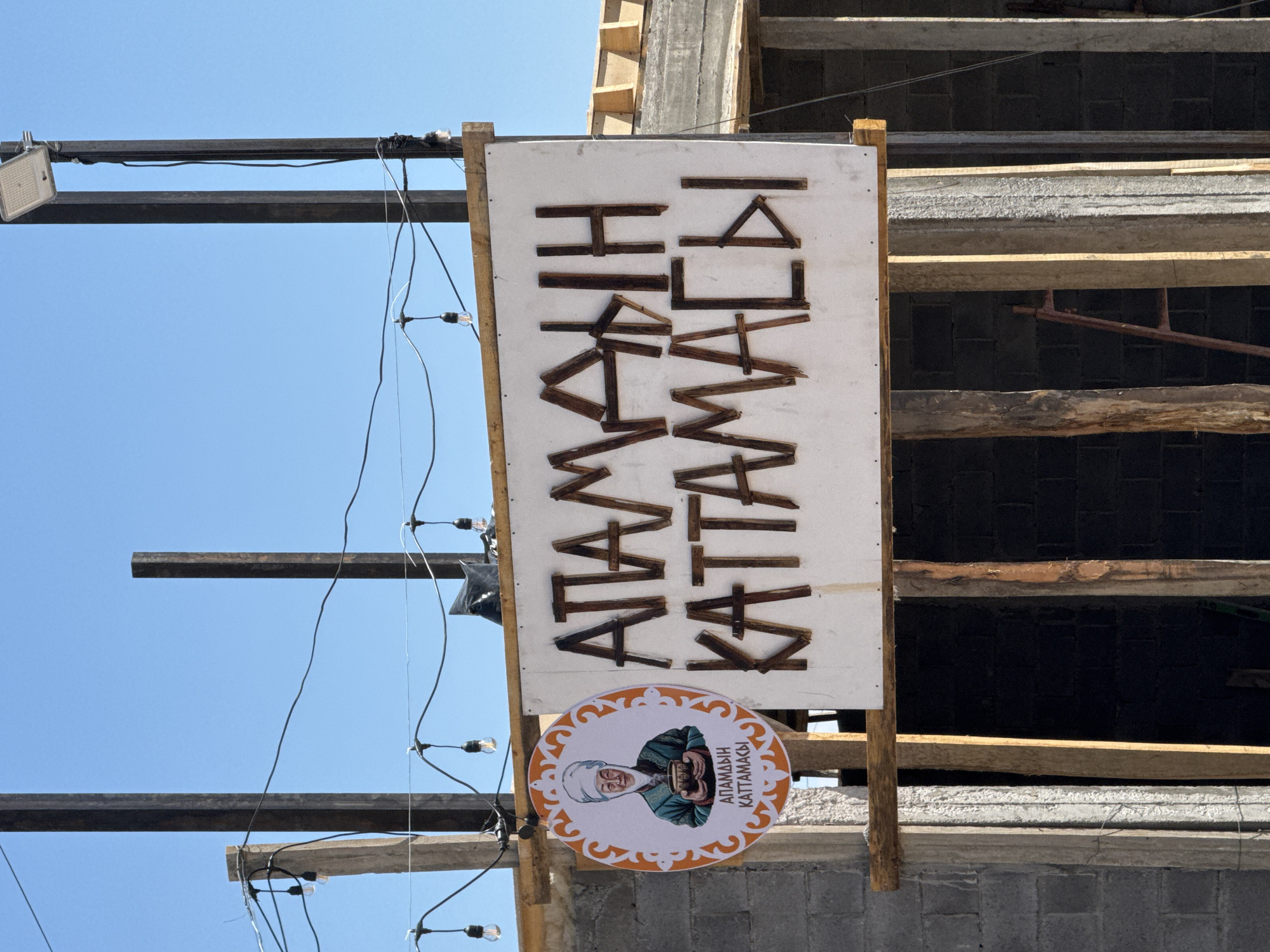 The image shows a sign for "ATAMDIN KATTAMASI" in Sary-Oy, Kyrgyzstan. The sign is a white rectangle with wooden letters. To the left of the sign is a circular logo with an image of a woman wearing a traditional headdress and holding a bowl. The sign is suspended by wires from metal poles, with decorative lights strung between them. In the background, a building is under construction, with visible concrete blocks and wooden beams. The sky is clear and blue.