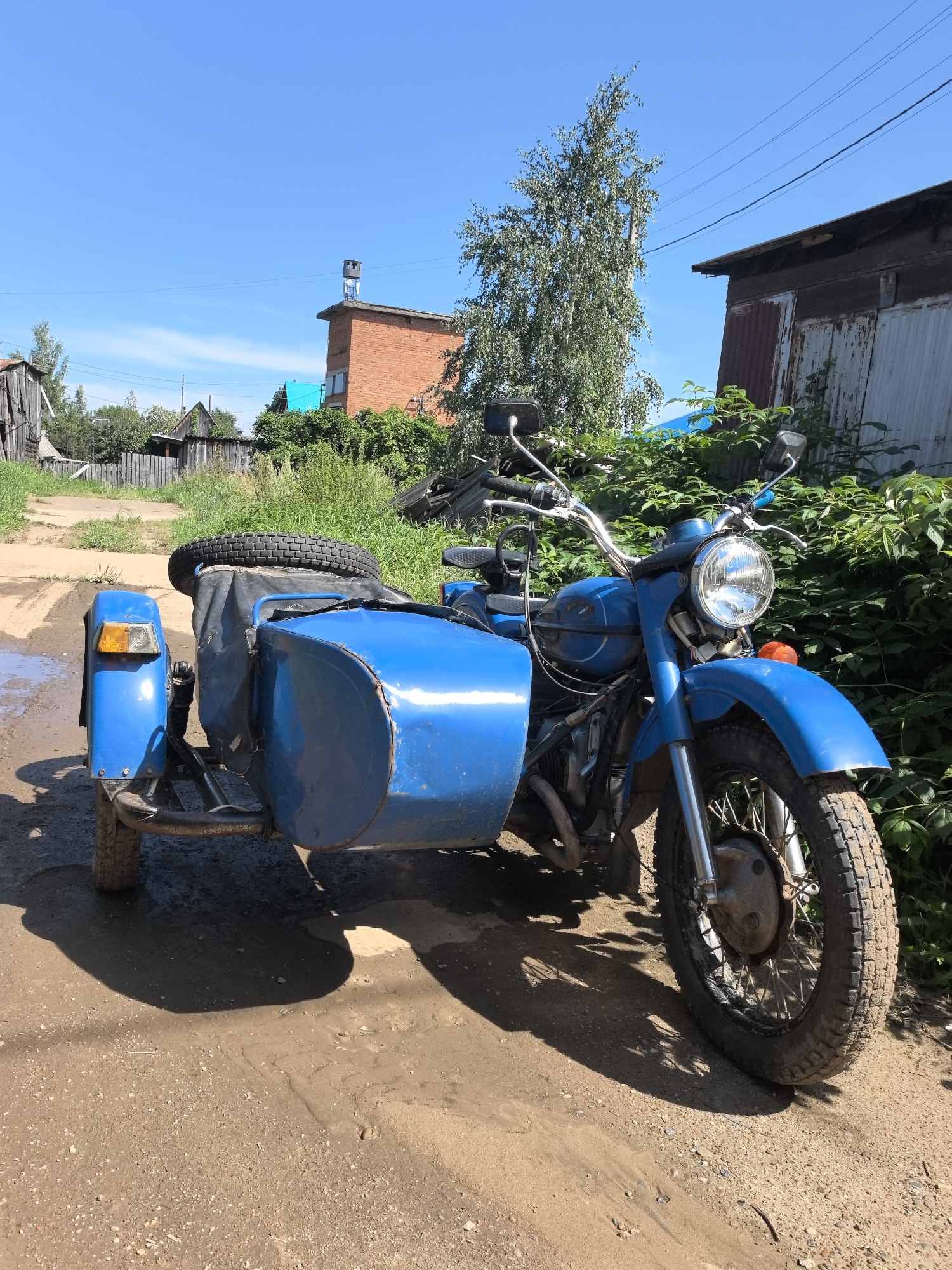A blue vintage motorcycle with a sidecar is parked on a dirt road in Noviy, Russia. The motorcycle has a spare tire strapped to the back of the sidecar. The scene is set on a sunny day with a clear blue sky. In the background, there are several wooden houses and a brick building, along with overgrown green vegetation. Power lines are visible overhead. The ground is a mix of dirt and gravel, with some areas showing signs of moisture.