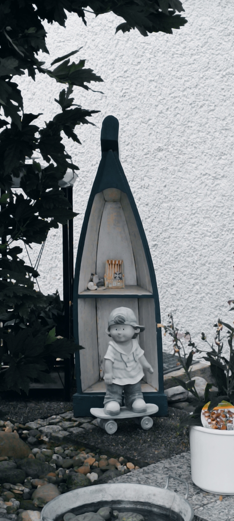 The image shows a garden scene in Waiblingen, Germany, with a decorative boat-shaped shelf mounted on a textured white wall. The shelf is dark blue with a light interior and has two shelves. On the top shelf, there are small objects, including what appears to be a miniature cuckoo clock with a tiny bird. On the lower shelf, a ceramic statue of a boy wearing a cap, t-shirt, and shorts stands on a skateboard. The statue is approximately 2 feet tall. The ground in front of the shelf is covered with small stones and gravel. To the left of the shelf, dark green foliage from a tree hangs into the frame. To the right of the shelf, small plants with light-colored flowers grow. In the foreground, a large, shallow metal tub with a handle is visible, containing some dark objects. A white pot is also present to the right of the tub. The overall lighting suggests it is daytime, with a slightly overcast or diffused light.