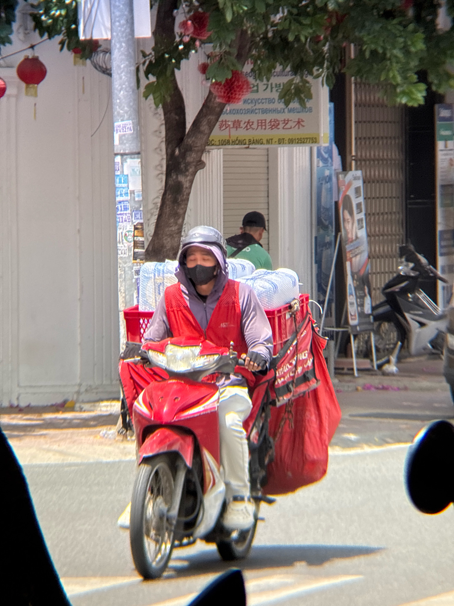 A man wearing a black mask, a gray helmet, and a red vest over a purple hoodie rides a red motorcycle. He is carrying a large red bag on the back of his motorcycle. The motorcycle is in motion on a paved road. In the background, there are buildings, a tree with red flowers, and other motorcycles parked. The scene appears to be in a city in Vietnam, likely Nha Trang, given the context. The image is a medium shot, capturing the man from the waist up.