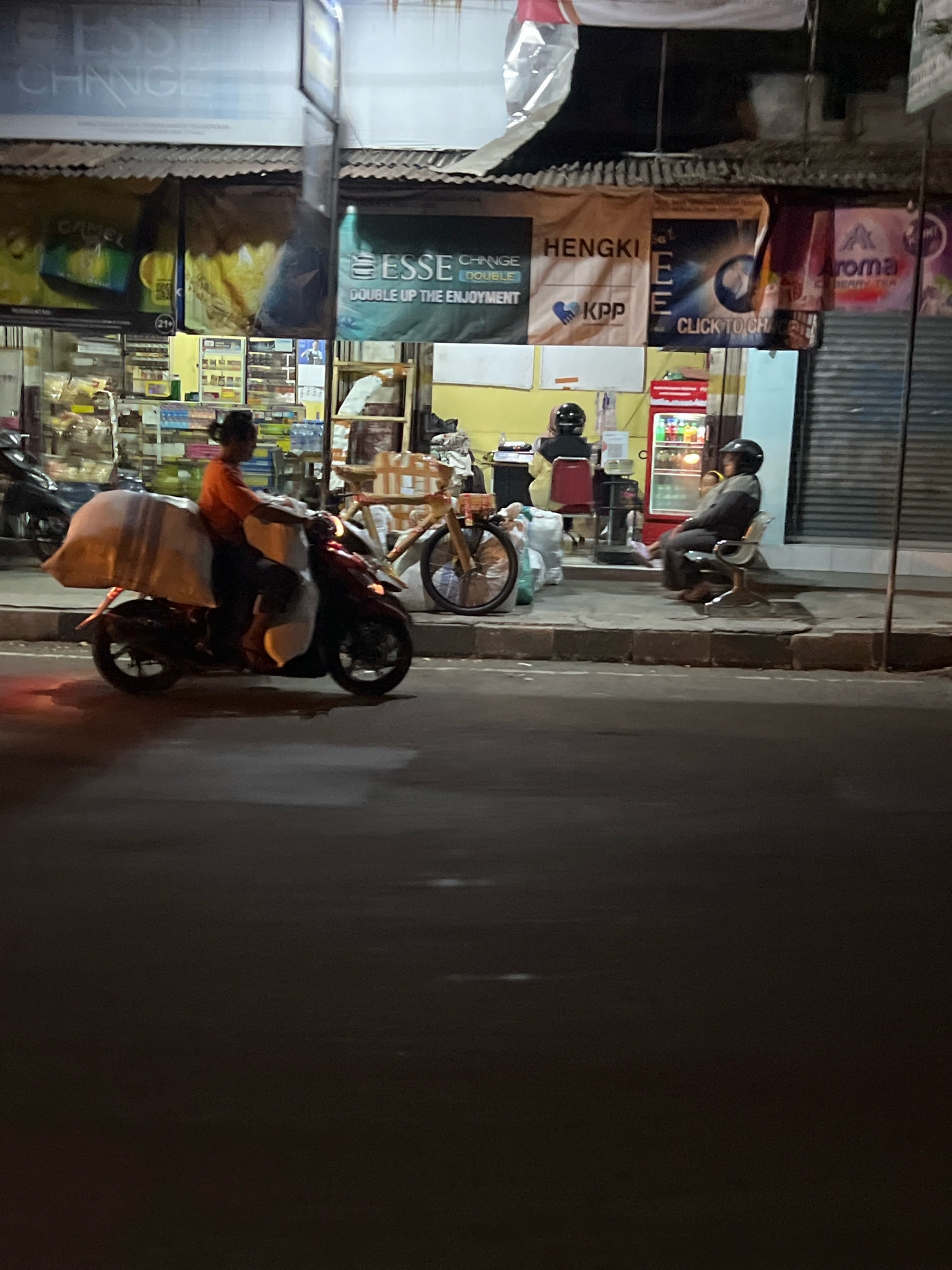A nighttime street scene in Sleman, Indonesia, features a person riding a motorcycle carrying a large, white, covered load. The motorcycle is red and appears to be a scooter. In the background, a shopfront displays various advertisements. One banner reads "ESSE CHANGE DOUBLE UP THE ENJOYMENT." Another banner says "HENGKI KPP." A third banner displays an "Aroma" logo with "BERRY TEA" underneath. A red refrigerator is visible inside the shop. Two individuals are seated on chairs outside the shop. One person, wearing a dark jacket and helmet, sits on a metallic chair. Another person, with a light-colored garment and a helmet, is seated further back. The scene is illuminated by artificial lighting, creating shadows on the dark asphalt road.