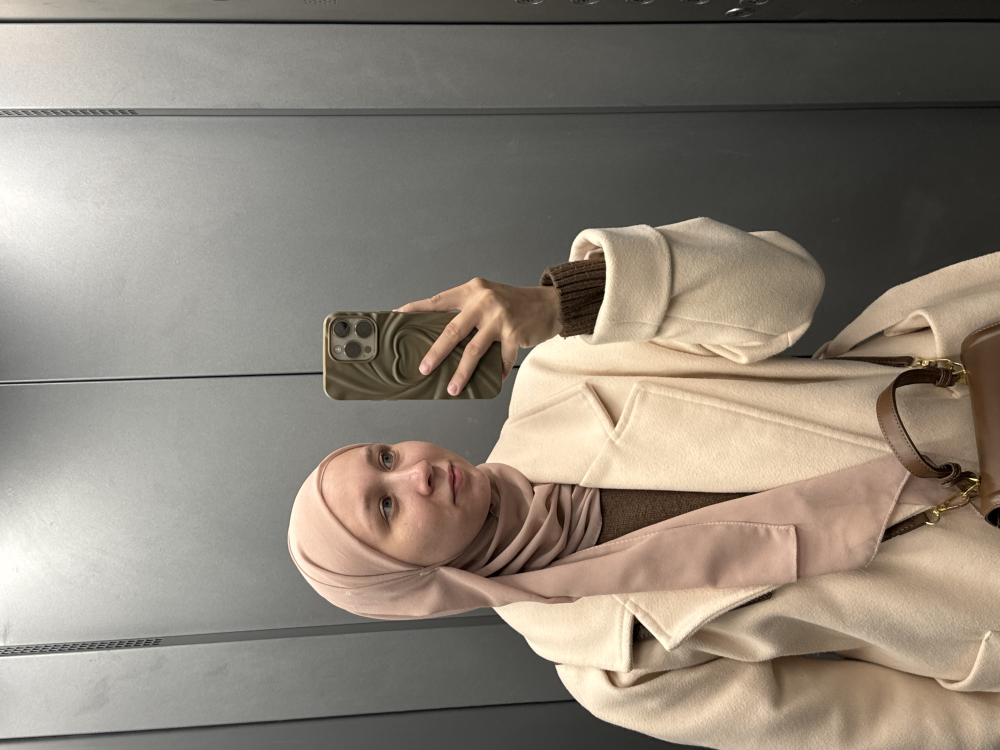 A woman wearing a hijab and light-colored coat is taking a selfie in what appears to be an elevator. She is holding a smartphone with a decorative case. The interior of the elevator is lined with gray metal panels. There are no identifying features suggesting the location of Chaikovsky, Russia.