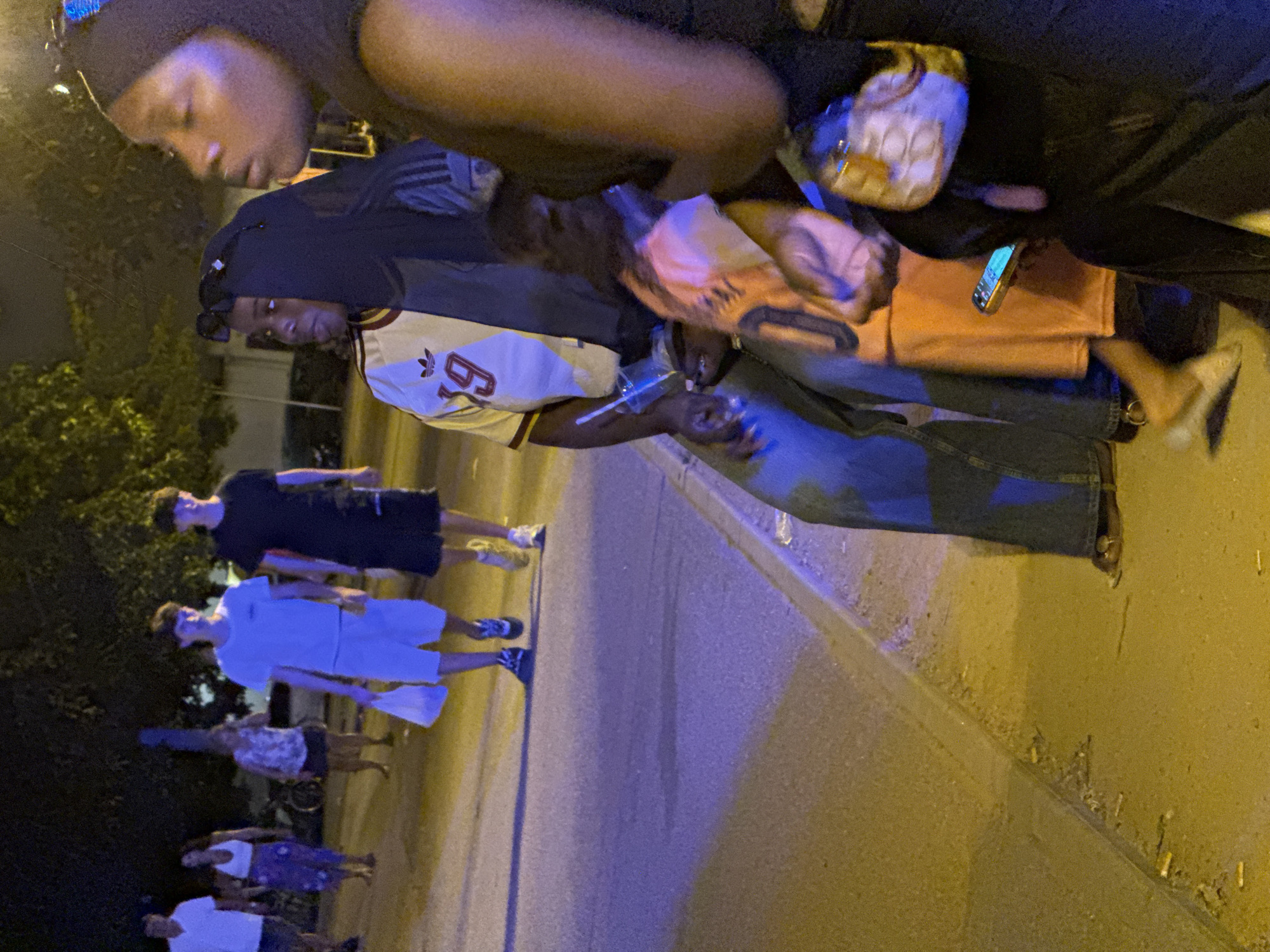 A group of people are walking along a sidewalk at night, with some sitting on the curb. The scene appears to be in Blanes, Spain, with an alleyway or street to the left and trees and buildings in the background. The lighting is dim, with a blue and yellow cast from streetlights. One woman is wearing a headscarf and a distinctive t-shirt with a split design, one half white with "39" in red, and the other half dark. She is holding a plastic cup with a straw. Another woman, also wearing a headscarf, is partially visible to her right. In the background, several individuals are walking away from the camera, some carrying bags. The ground is littered with cigarette butts.