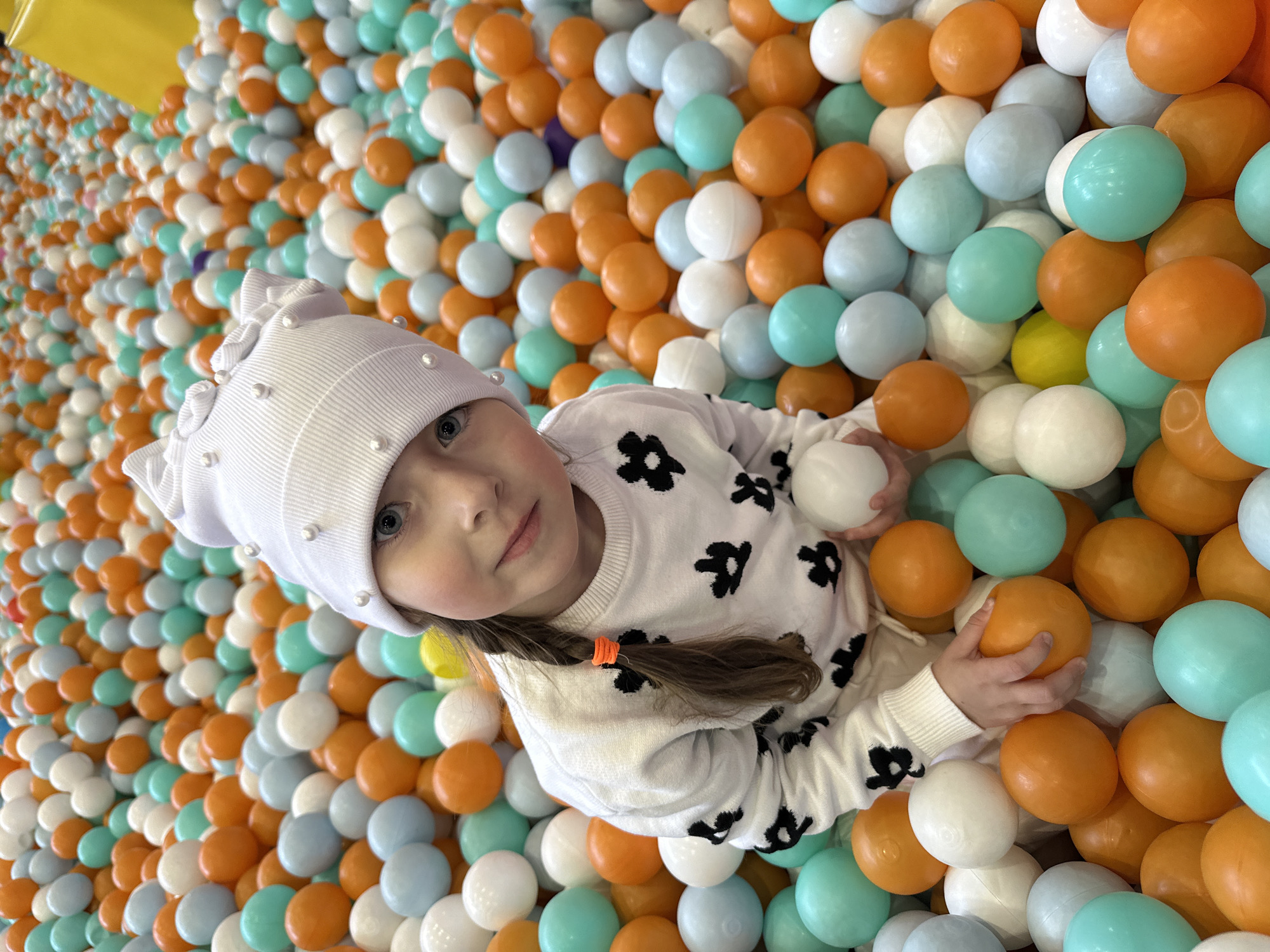An overhead, angled shot captures a young girl playing in a vibrant ball pit, likely within an indoor play area in Sochi, Russia. The girl, with fair skin and striking blue eyes, looks directly at the camera with a calm, engaged expression. Her light brown hair is tied into a braid with a bright orange elastic. She wears a white knitted hat embellished with small white pearls and a white long-sleeved sweater featuring scattered black floral or abstract patterns.
She is immersed in a large volume of plastic balls, predominantly colored in bright orange, white, light blue, and teal/mint green. A few yellow balls and one dark purple ball add slight variations to the colorful array. Her hands are visible, gently holding some of the orange balls. The extensive ball pit fills the entire frame, with a glimpse of a yellow wall or boundary visible in the top left corner, indicating the perimeter of the play area. The scene conveys a cheerful and playful atmosphere.