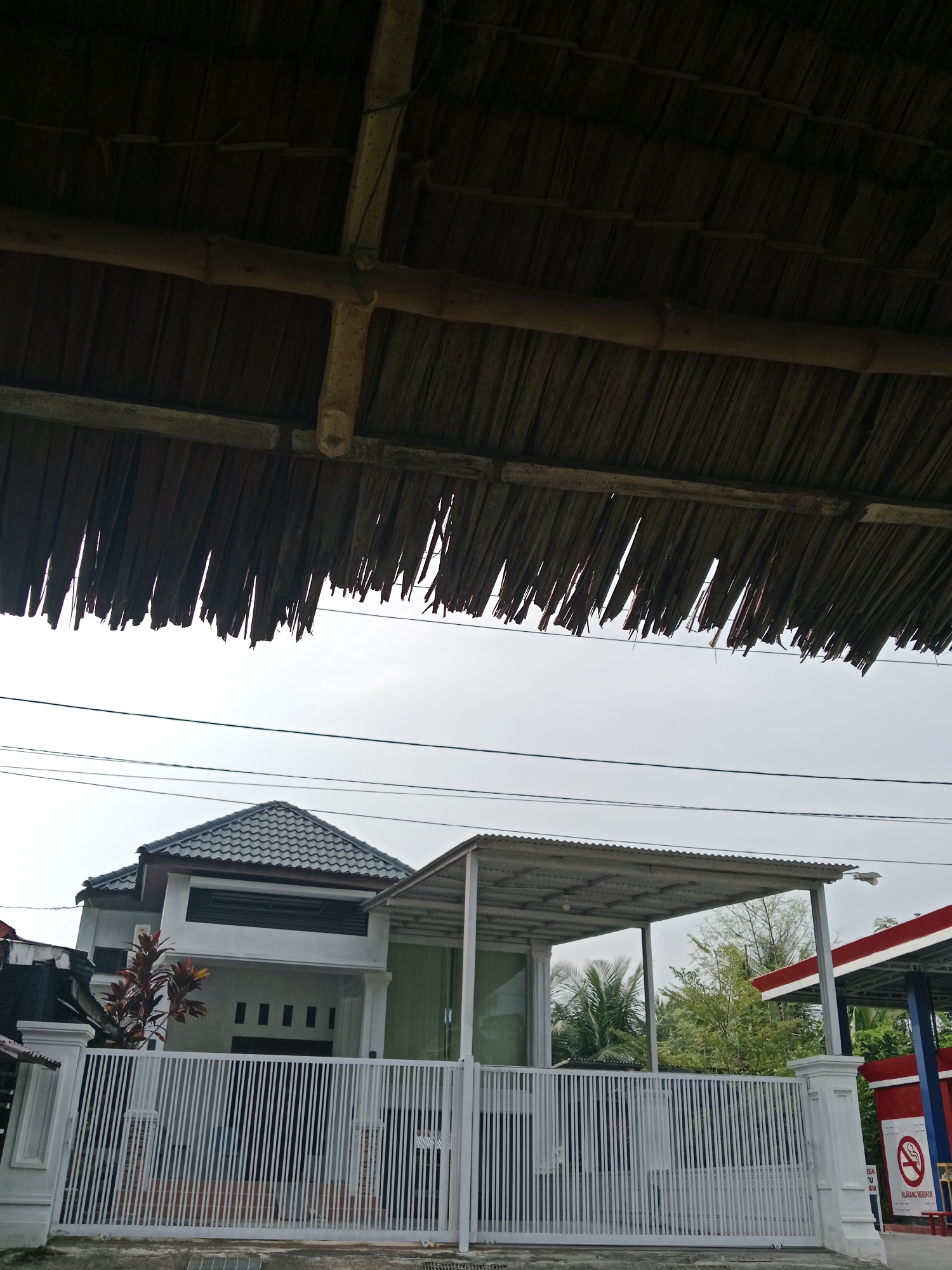 The image shows a view from under a thatched roof in Meunasahara, Indonesia. In the foreground, the bottom edge of the roof is visible, composed of dark, dried plant material supported by bamboo poles. Below the roof, a modern, multi-story house with a grey tiled roof and white walls stands behind a white metal fence. The house has large windows and several smaller rectangular openings on the lower level. To the right of the house, a structure with a corrugated metal roof, possibly a porch or carport, is visible. Further to the right, part of a gas station with red and blue accents and a "no smoking" sign is present. Power lines are strung across the sky in the middle ground. Trees, including palm trees, are visible behind the house and the gas station, suggesting a tropical environment. The sky is overcast, indicating a cloudy day. There are no people or animals visible in the image.