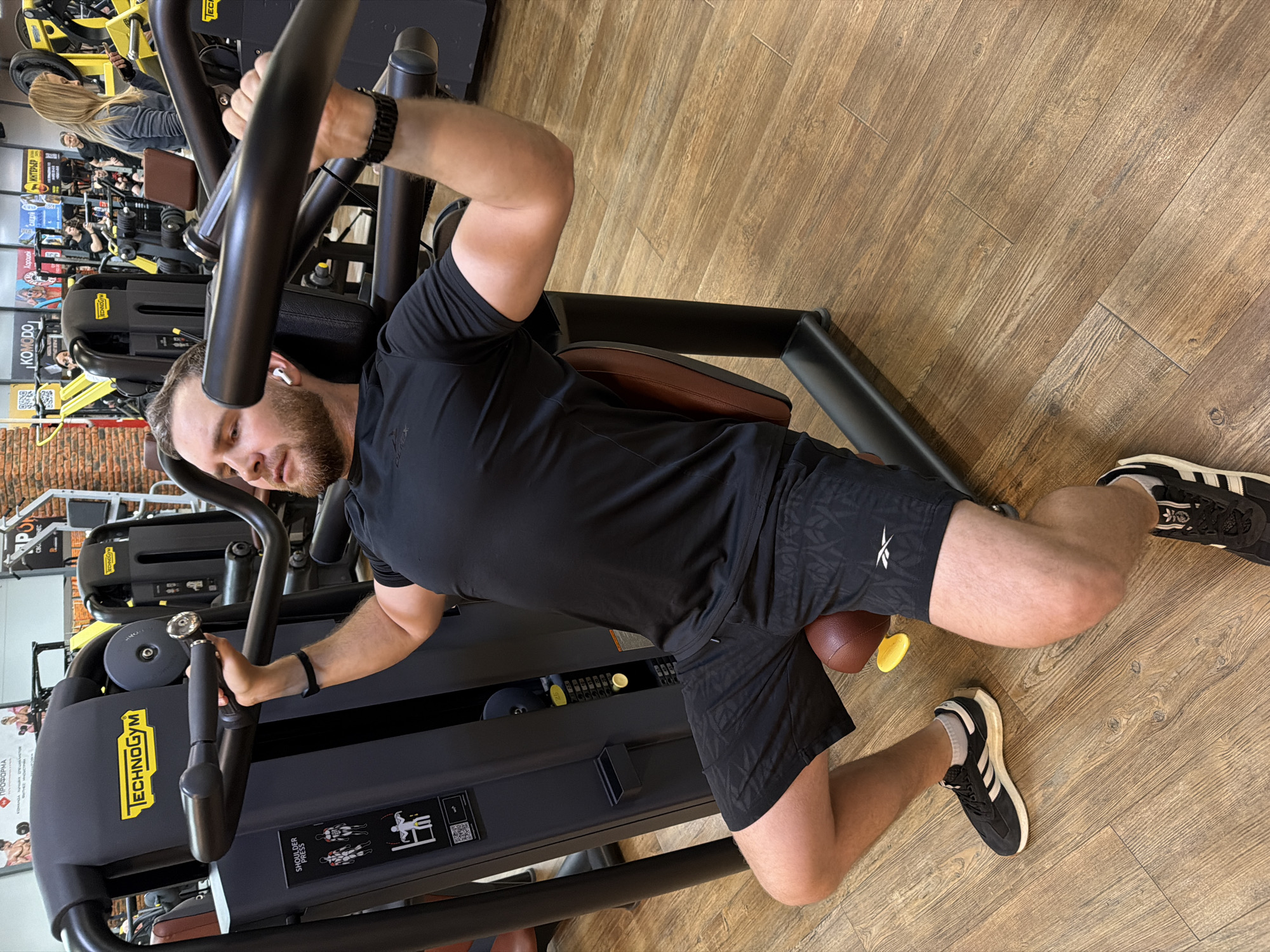 A man is using a Technogym shoulder press machine in a gym in Chaikovsky, Russia. He is wearing black athletic shorts, a black t-shirt, and black Adidas sneakers with white stripes. The gym has a wood-look floor. Other exercise equipment and gym patrons are visible in the background.