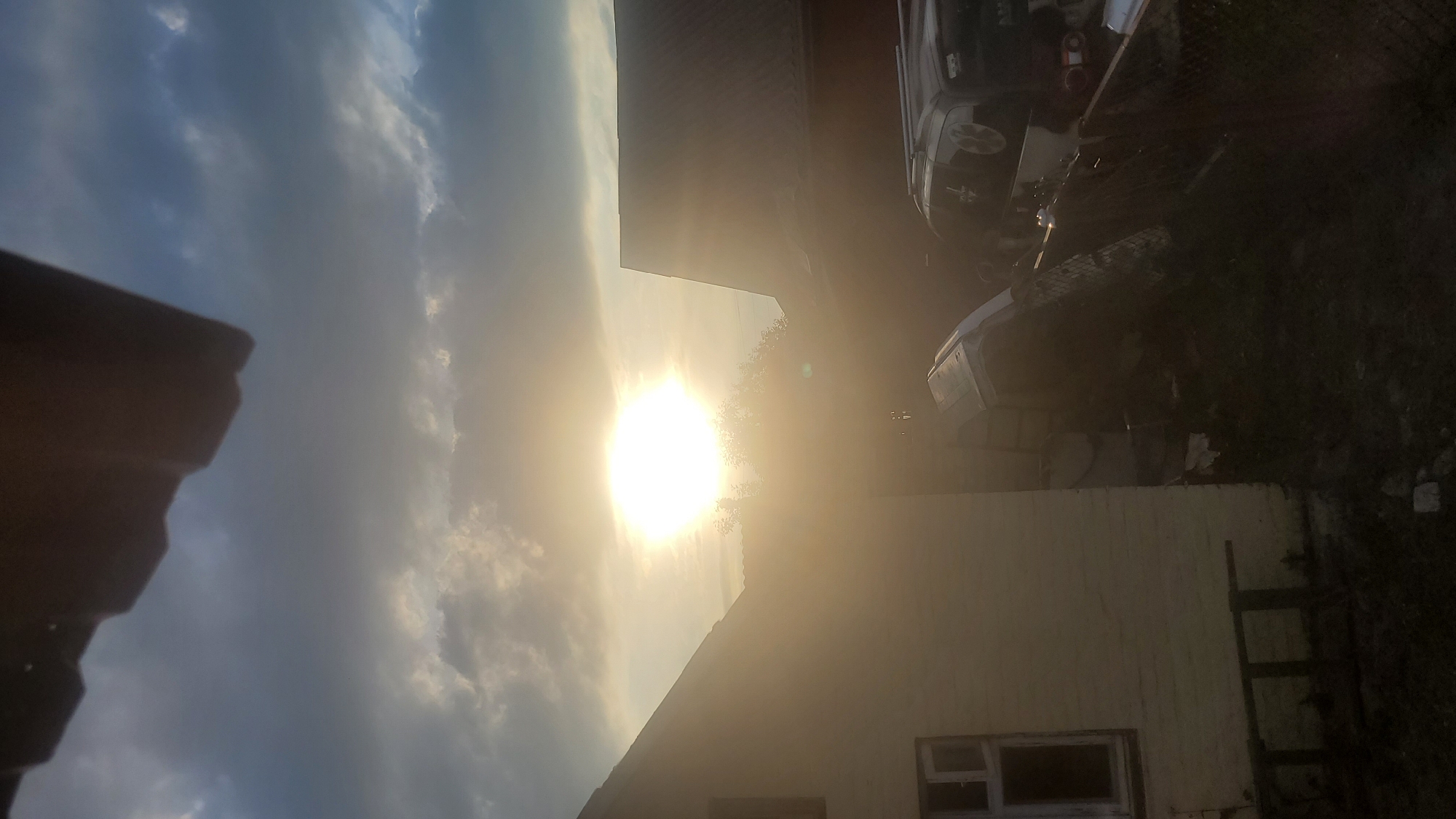 The image shows an outdoor scene, likely a residential backyard, under a dramatic sky with a bright sun partially obscured by clouds. The sun is positioned in the upper middle of the frame, emitting a strong glare. In the foreground, to the left, is the side of a light-colored building, possibly a house or shed, with a visible window frame. To the right of the building, a dark-colored vehicle with roof racks is parked. In the center of the image, lying on its side and partially obscured by shadows and the glare, is what appears to be a white vehicle or large object. A wire fence separates the foreground from the background on the right side. The overall lighting suggests either sunrise or sunset, with a hazy, warm glow. The scene is devoid of any visible people or animals. The background consists of trees and the roofs of other buildings. The location is specified as Unknown, Unknown.