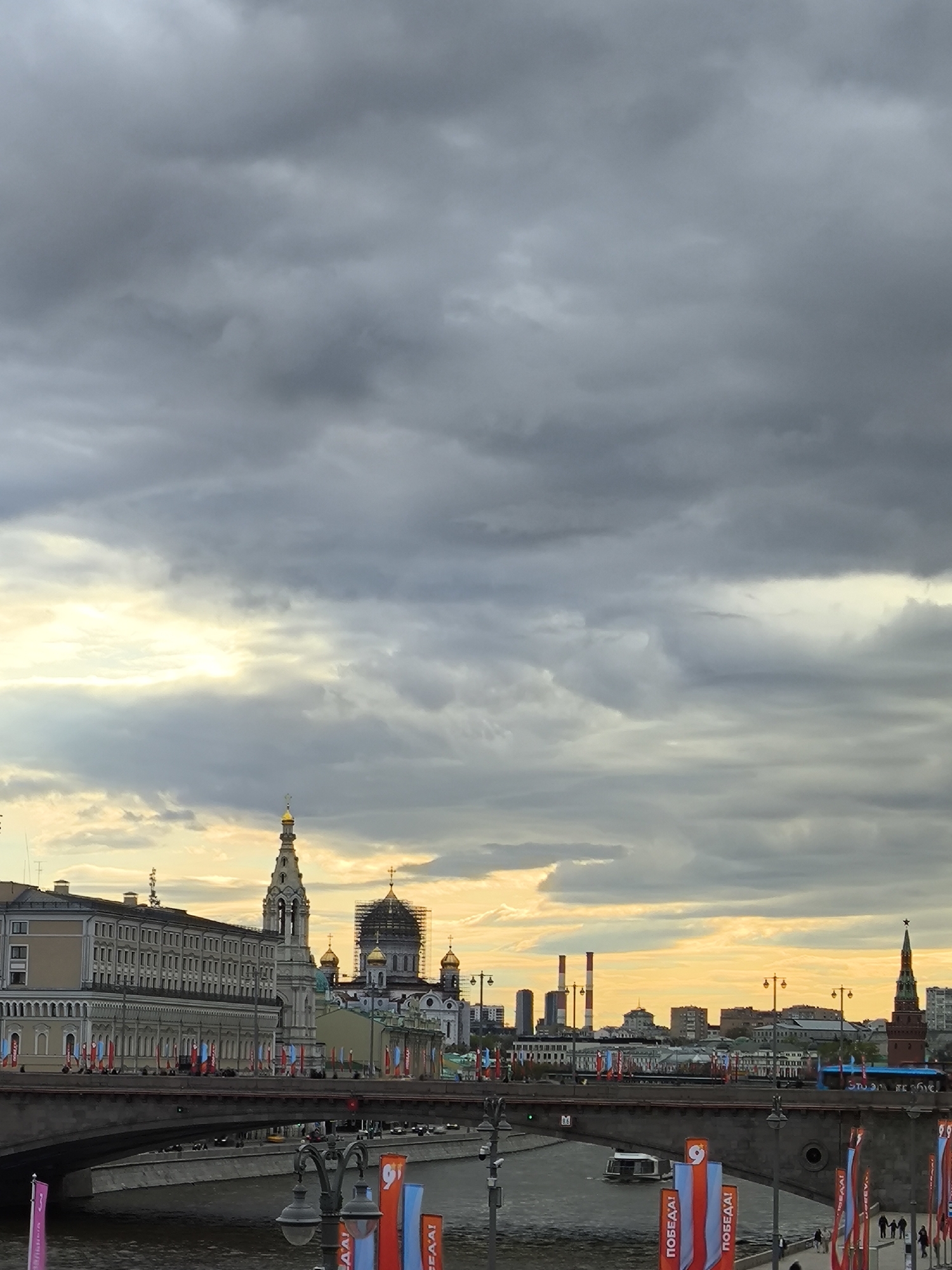 The image shows a cityscape in Moscow, Russia, under a cloudy sky. A bridge spans a river in the foreground. Along the river and bridge are red, white, and blue banners with Cyrillic text. In the midground, there's a building with a yellow façade, a bell tower with a golden dome, and the Cathedral of Christ the Savior undergoing some sort of construction or renovation. Other buildings and smokestacks are visible in the distance. The sky is dominated by thick, grey clouds with hints of sunlight peeking through.