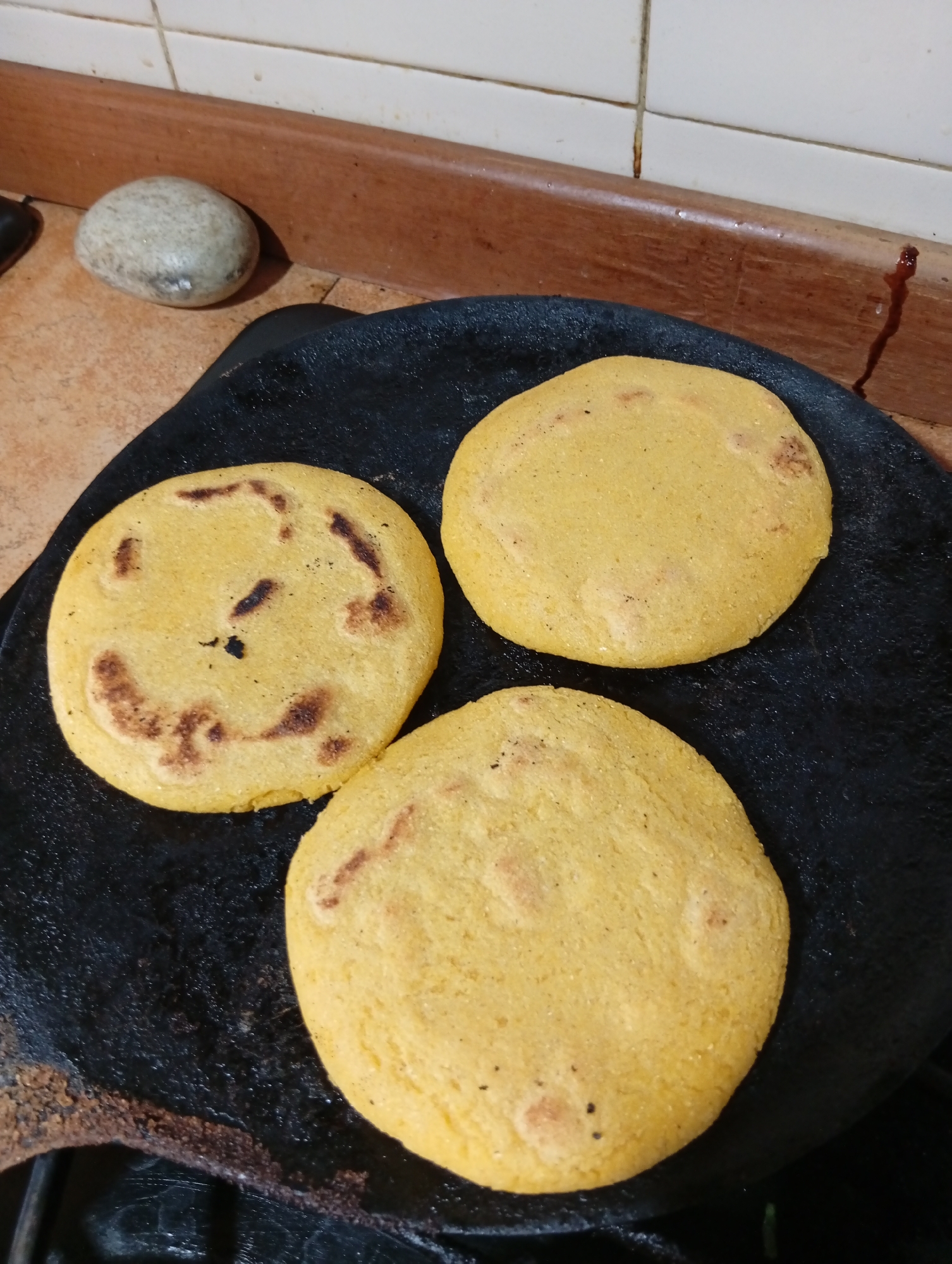 Three golden corn arepas are cooking on a black, flat griddle. The arepas are circular and have some browning and char marks, indicating they are being toasted. The griddle is placed on a countertop with a tiled backsplash and a wooden trim above it. A smooth, grey stone rests on the countertop to the left of the griddle. The overall environment appears to be a kitchen. The specific location of San Antonio de Los Altos, Venezuela, is not discernible from the visual information present in the image.