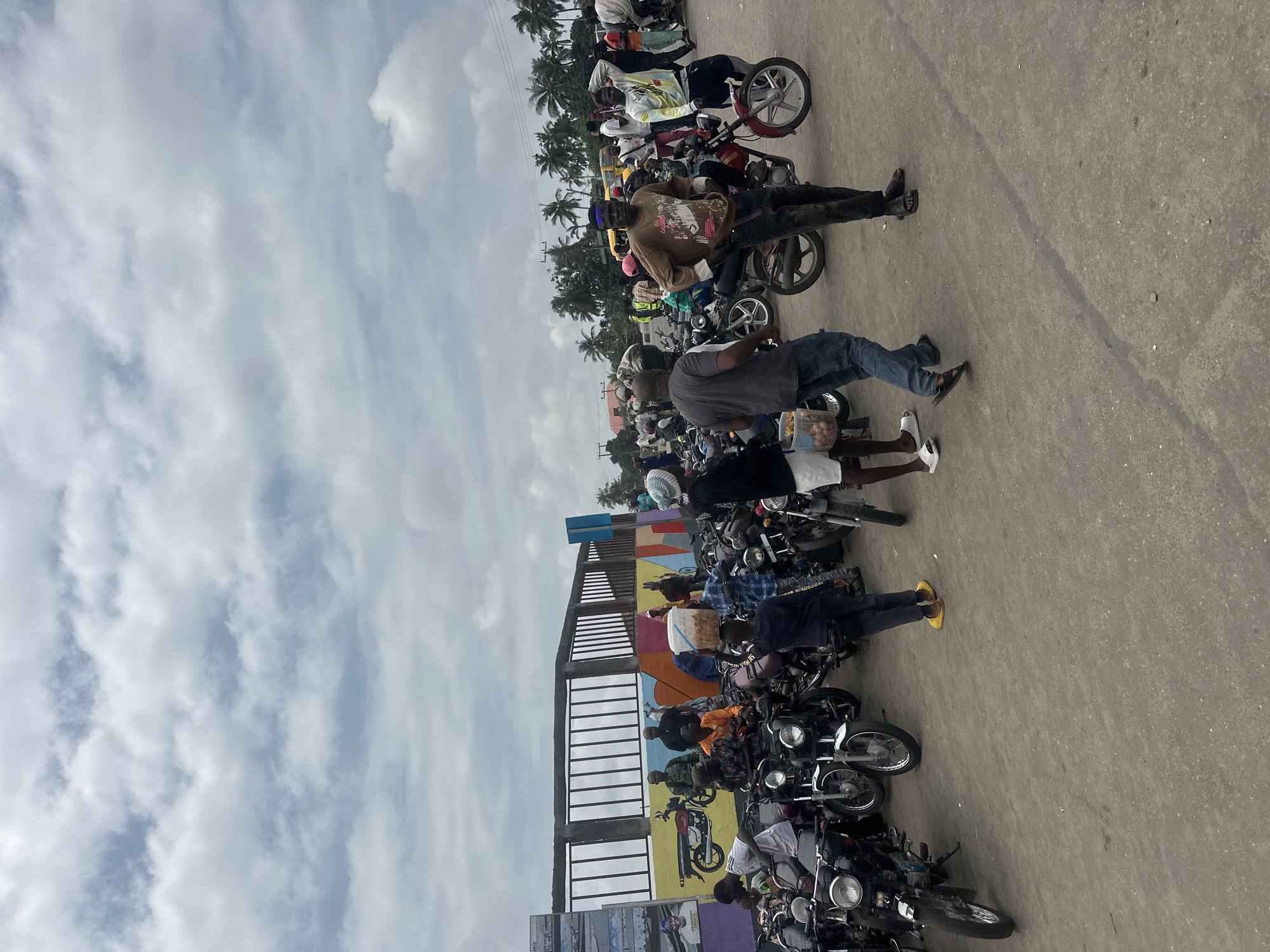 The image depicts a busy outdoor scene in Badagry, Nigeria, characterized by a large gathering of motorcycles and people. The sky is overcast with scattered clouds. In the background, a wall with a colorful mural featuring abstract designs and a drawing of a motorcycle is visible, behind which are palm trees and buildings. A fence with vertical bars runs along the top of the mural.
The foreground is dominated by numerous motorcycles parked closely together, suggesting a transport hub or a marketplace. Several individuals are present, some standing, some seated on the motorcycles, and some walking. They are casually dressed in various attire, including t-shirts, shirts, trousers, and shorts. One person is wearing a brown t-shirt and dark trousers, with sunglasses perched on their head. Another person in the foreground is wearing a blue and white checkered shirt, dark trousers, and carrying a bucket on their head filled with produce. A person in a black t-shirt and dark pants is standing with one foot on a motorcycle. A woman in a light blue headscarf and a blue top is seen near the bucket carrier.
The ground is unpaved, appearing to be a dirt or gravel surface. The overall atmosphere suggests a lively, everyday scene in a Nigerian city, likely a place where people use motorcycles for transportation or commerce. The presence of the mural and the specific types of motorcycles might indicate a particular area within Badagry, possibly near a historical site or a main road.