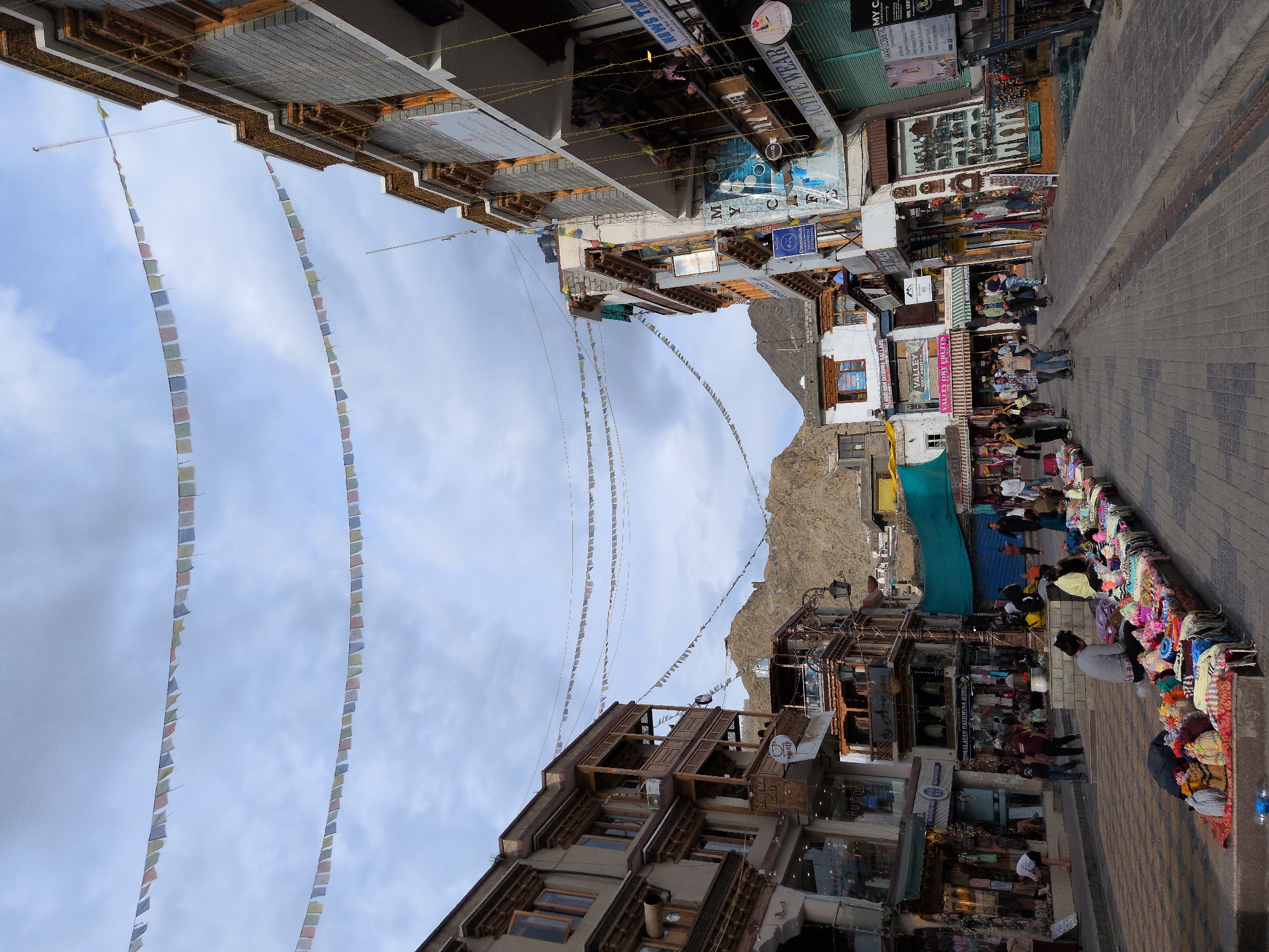 The image depicts a bustling street scene in Leh, India, with prayer flags strung across the sky. The street is lined with multi-story buildings, many of which house shops and cafes. The architecture appears to be a blend of traditional Ladakhi and more modern styles, with intricately carved wooden balconies and stone facades.
In the foreground, a street vendor displays an array of colorful textiles and handicrafts, including woolen shawls, scarves, and hats. Several people are browsing the merchandise or walking along the street. The background reveals the majestic, barren mountains that surround Leh, hinting at the city's high-altitude location. The sky is overcast with a mix of clouds and patches of blue.