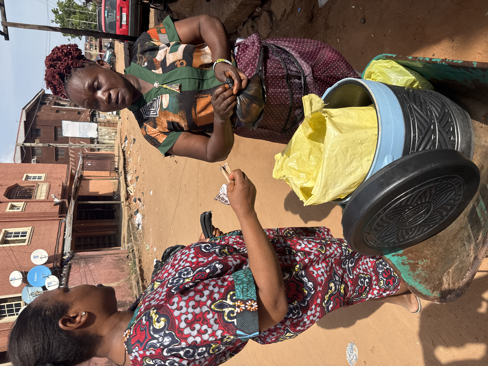 This image captures a vibrant street scene in Onitsha, Nigeria, showing a transaction between two women under clear daylight.
The primary subjects are two women. The woman in the upper part of the frame, likely a vendor, wears a green and patterned short-sleeved top with red braided hair. She is actively preparing an item, securing a black plastic bag that appears to hold food or goods, her hands focused on the task. She also wears a purple polka-dotted skirt or wrapper and a black bracelet with white text, possibly "NEW GOD".
Facing her is a customer, dressed in a striking red, black, and yellow patterned top. She holds money, folded neatly, in her right hand, ready to complete the purchase, and a small black pouch or wallet in her left. Her gaze seems directed towards the vendor or the item being prepared.
The activity taking place is a typical street vending transaction. The vendor is packaging an item from a large, light-blue banded cylindrical container, which is partially filled with yellow plastic bags, presumably holding other items for sale. Behind this container is a green, rectangular bin also containing yellow bags. The black lid of the cylindrical container has "GERMAN" embossed on it.
The setting is an outdoor, bustling environment, indicated by the dusty ground, various buildings in the background, and a red car visible further down the street with a license plate partially visible ("116-SM"). On one of the buildings, several satellite dishes, one clearly marked "DSTV", are mounted. A sturdy wooden utility pole stands on the left. The bright sunlight suggests a warm day, casting clear shadows. The overall scene reflects a common daily interaction in an urban or semi-urban area of Nigeria.