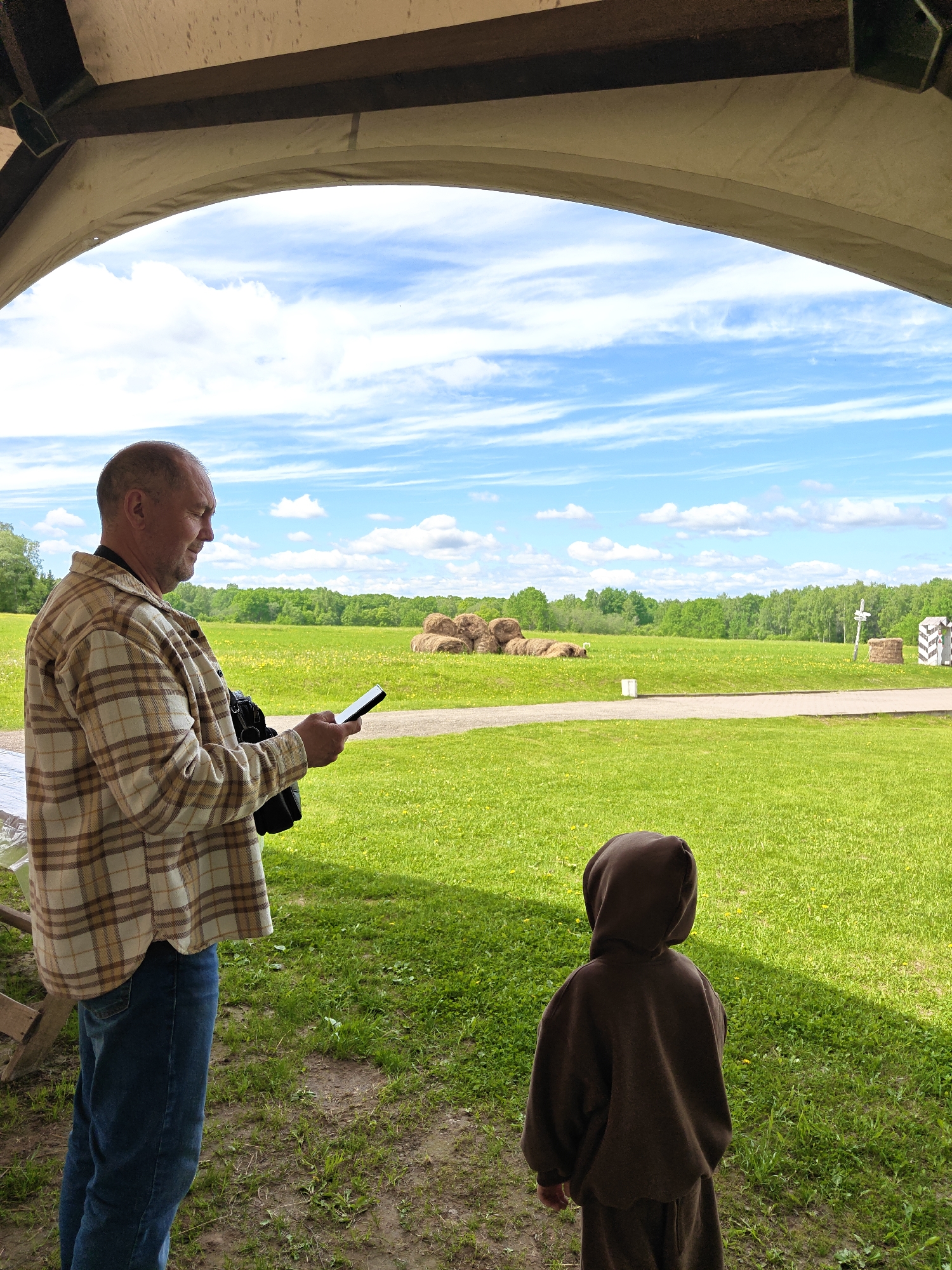The image shows a man and a child standing under a light-colored awning in an outdoor setting. The man, with a shaved head, is wearing a plaid shirt and jeans and appears to be looking at a phone. A black camera bag is visible on his shoulder. The child, seen from behind, is wearing a brown hooded sweatshirt and pants.
The background features a grassy field with trees in the distance and bales of hay or straw. A decorative signpost is also present. The sky is partly cloudy. The location is Borodino, Russia.