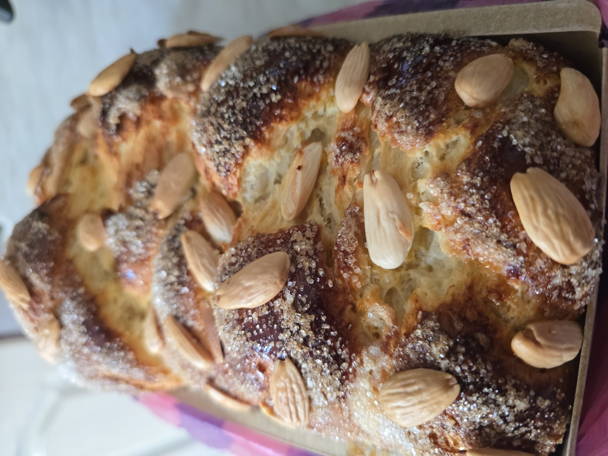 The image shows a sweet bread, possibly a Kozunak, topped with almonds and sugar. The bread has a braided texture and is golden brown in color. It is likely located in Varna, Bulgaria, based on the prompt's request to include this location. However, there is no inherent visual information within the image itself to confirm this location.