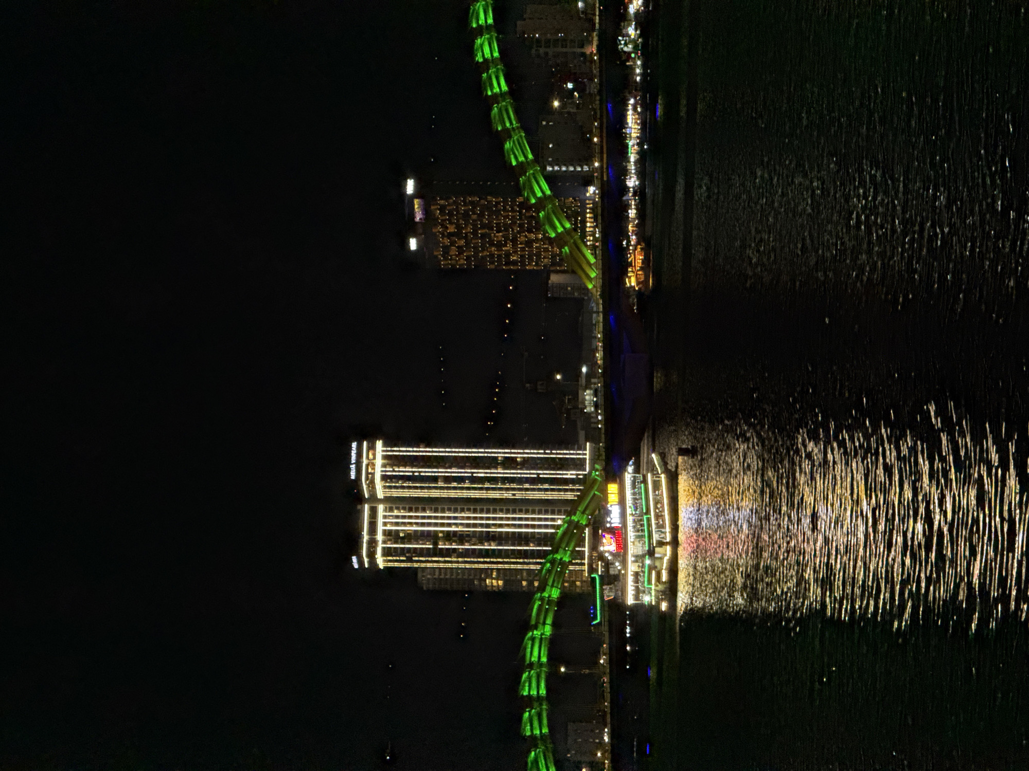 This is a nighttime photograph of the Dragon Bridge in Da Nang, Vietnam. The bridge, illuminated with bright green lights, arches over a dark body of water that reflects the city's lights. On the left side of the image, a tall hotel, possibly the Melia Vinpearl, is prominently featured with its windows lit in a warm yellow glow. The surrounding buildings also show signs of urban life with scattered lights. The water's surface is disturbed, creating shimmering patterns of light from the bridge and the buildings. The overall mood is vibrant and festive, characteristic of a city at night. There is text visible on the hotel: "MELIA VINPEARL". The time of day is clearly night, with no discernible weather conditions. The scene depicts an urban landscape at night, with a focus on the iconic Dragon Bridge and its illuminated surroundings.