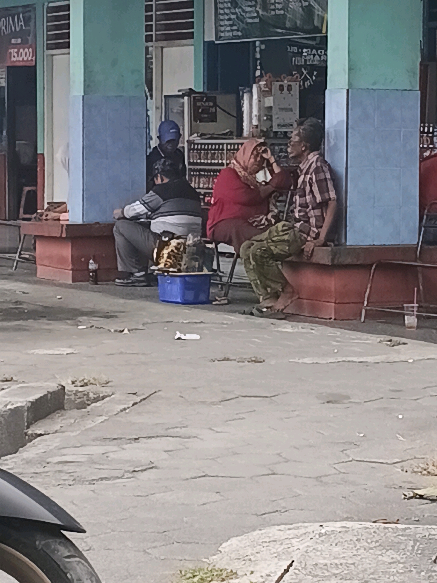 In Kuripan, Indonesia, a man in a plaid shirt and patterned pants sits on a red platform next to a blue tiled wall. A woman wearing a red hijab and a dark colored top is seated beside him, applying makeup to his face with her right hand. Another person, partially visible, sits behind them. In front of them is a blue plastic container filled with items. To the left of the platform, a sign with the text "PRIMA 15.000" is visible on a blue wall. The ground is paved with a cracked surface, and a motorcycle fender is visible in the foreground.