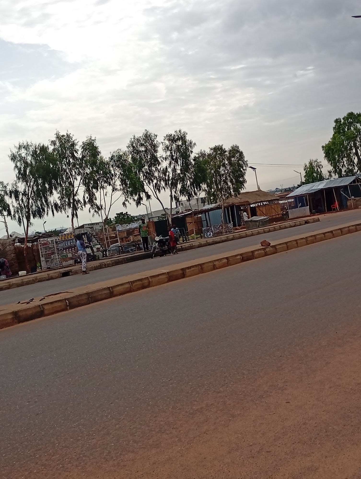 The image shows a street scene in Wuro Dole, Nigeria, with a paved road in the foreground and a sidewalk lined with small shops and trees on the right. A few people are visible, including a woman standing on the sidewalk, a man in a green shirt, and two individuals near a motorcycle. The shops appear to be selling various goods, with some items displayed outside. The sky is overcast, suggesting a cloudy day. The overall impression is of a typical day in a Nigerian town, with economic activity and community interaction taking place along the street.
