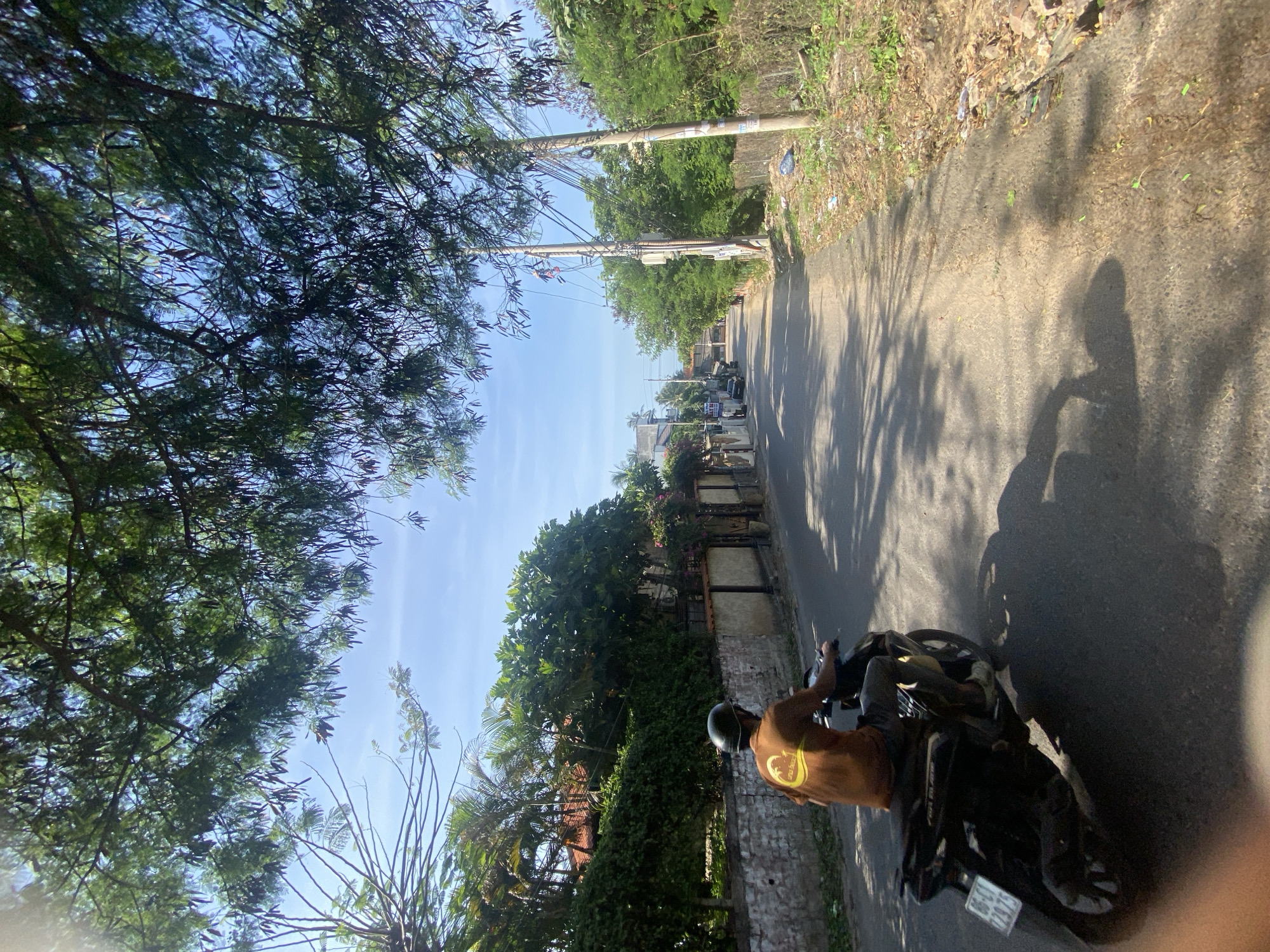 A person wearing a helmet and a brown t-shirt with a circular logo is riding a dark-colored scooter down a paved road in Mũi Né, Vietnam. The scooter has a license plate with Vietnamese characters and numbers. The road is lined with houses, lush green trees, and vegetation. Electrical poles and wires are visible on the right side of the road. The sky is blue with some clouds. The sun is shining, casting shadows on the road from the trees and the scooter. The overall scene depicts a typical sunny day in a residential area of Mũi Né.