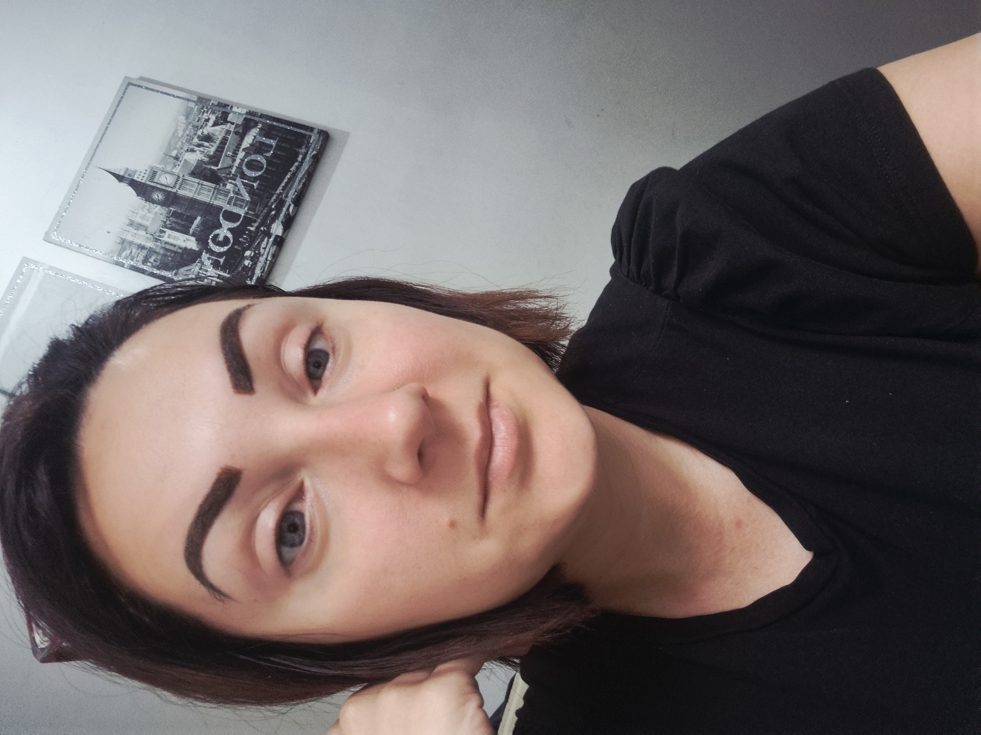 A young woman with dark brown hair and fair skin is pictured in a close-up shot. She has on a black V-neck t-shirt. Her left hand is supporting her head. In the background, a framed print of Big Ben in London is visible on a light gray wall. The image does not provide any information about the location of Sandwell, United Kingdom.