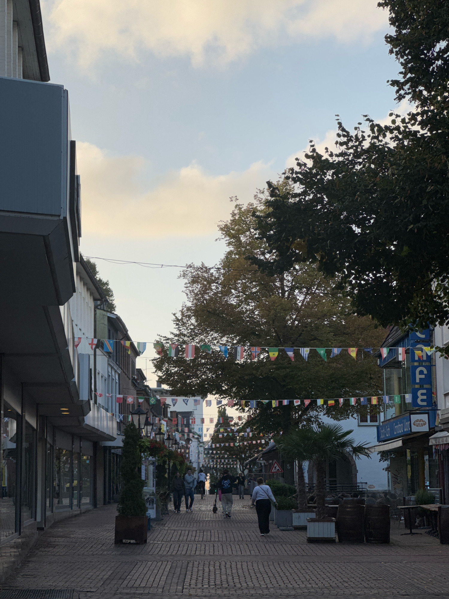 The image depicts a pedestrian street in Cleves, Germany, decorated with numerous international flags strung across the pathway. The street is paved with red brick and lined with buildings on both sides. On the left, a modern-looking building with large glass windows and a dark gray overhang dominates the foreground. On the right, a mix of storefronts and buildings with awnings and signage are visible, including a blue sign with white letters that appears to read "CAFÉ". Several people are walking along the street, some individually and others in small groups. Trees, including a palm tree, are planted in planters along the right side of the street. In the distance, the street recedes, suggesting a commercial or shopping area. The sky is partly cloudy, with a soft, diffused light, indicating it may be late afternoon or early evening. The overall atmosphere is calm and suggests a typical day in a European town.