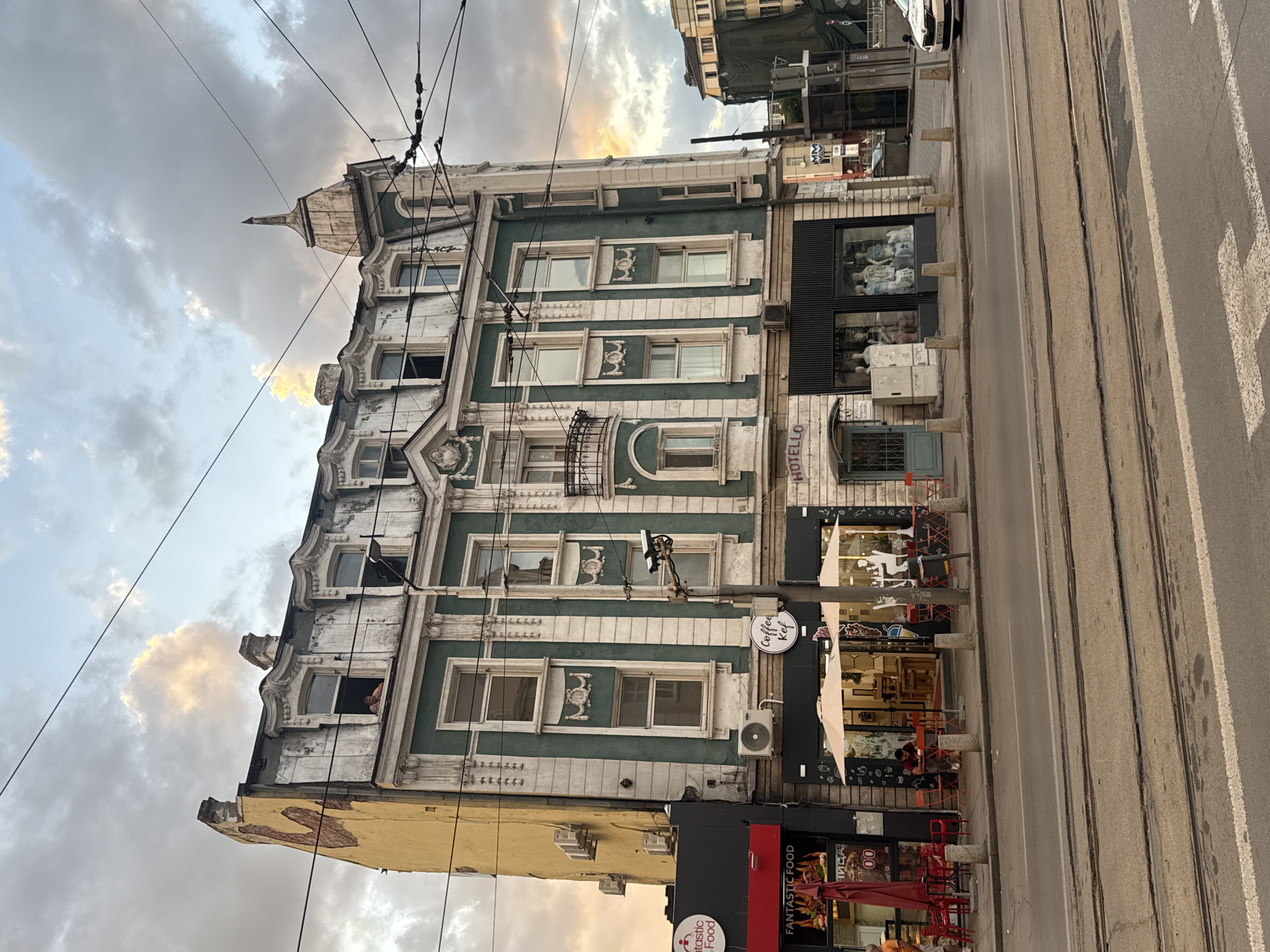 The image depicts a street scene in Sofia, Bulgaria, with a focus on an old, ornate building and the adjacent street. The building features a distinctive green facade with white decorative elements and numerous windows, some with elaborate detailing. Above the main facade, there is a pitched roof with a pointed turret. The building appears to be several stories tall.
At street level, the building houses several businesses. One storefront has a sign that reads "Coffee Kef." Another, with a black facade, displays mannequins in its windows, suggesting it's a clothing store. Signs for "FANTASTIC FOOD" are also visible. Red outdoor seating, likely for a cafe or restaurant, is arranged along the sidewalk.
Tram tracks run parallel to the sidewalk and the building, indicating this is likely a transit route. The time of day appears to be late afternoon or early evening, as indicated by the warm, diffused light and the color of the sky, which is filled with clouds, some catching the sunlight. The overall mood is somewhat atmospheric, with the architecture suggesting a historical European city. There are no overt activities or events taking place, beyond the typical functioning of shops and the presence of a few people seated outside. Overhead, power lines crisscross the sky, a common sight in urban environments.