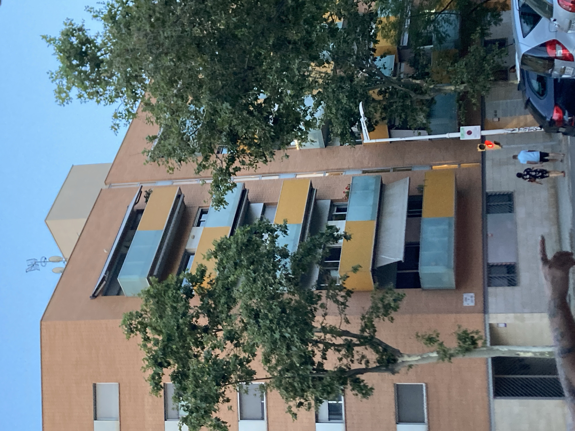 This image captures a street scene in Barcelona, Spain, featuring a multi-story building with a distinctive facade. The building is characterized by alternating panels of orange-brown brick and brightly colored sections in yellow and blue. These colored sections appear to be balconies or decorative elements, creating a visually striking pattern.
In the foreground, a large tree with green foliage partially obscures the view of the building, its branches extending across the frame. The sky is a clear, light blue, suggesting it is daytime, likely late afternoon or early evening given the lighting.
On the street level, a traffic light is visible, showing a red signal, indicating that vehicles are stopped. Two people are seen walking away from the camera, one wearing a light blue shirt and dark shorts, and the other in a dark patterned shirt and pants. To the right, parts of two cars are visible, one a dark blue vehicle and another silver car behind it.
A hand is visible in the lower right corner of the image, making a "rock on" gesture. This gesture, along with the casual attire of the people and the general urban setting, hints at a relaxed atmosphere. The presence of the traffic light and the building's architecture suggest a residential or mixed-use urban environment. No legible text is visible in the image.