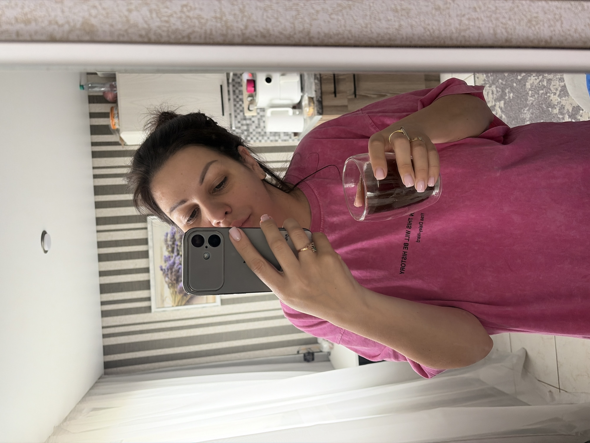 A woman with dark hair tied back in a bun is taking a mirror selfie in what appears to be a kitchen. She is wearing a bright pink t-shirt and holding a clear glass filled with a dark liquid, possibly coffee, in her right hand. Her left hand is holding a smartphone, capturing the reflection. She is also wearing two rings on her left hand. The background shows striped wallpaper, a framed picture of lavender on the wall, and kitchen cabinets with appliances. The presence of a water bottle on a shelf suggests a domestic setting. The location is not specified in the image or context provided.