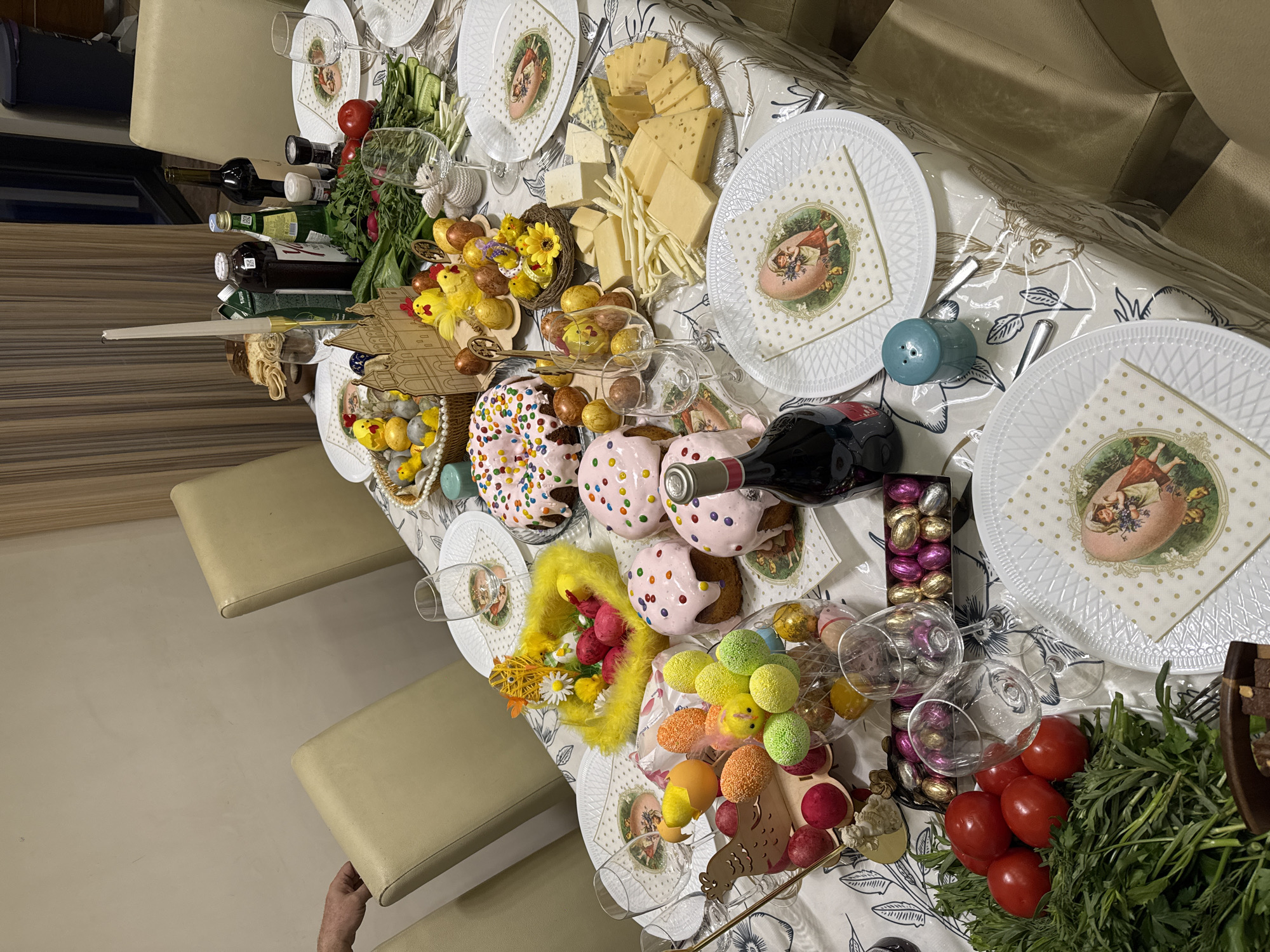 The image depicts a table laden with food and decorations, suggestive of an Easter celebration. The table is covered with a tablecloth featuring floral patterns. The setting appears to be indoors, perhaps in a private home.
Various dishes and items are visible on the table including decorated Easter cakes, a variety of cheeses, tomatoes, greens, and a selection of wines. There are also decorative elements, such as Easter eggs, figurines, and floral arrangements. The table is set for multiple diners with plates, napkins, glasses, and cutlery.
While the image provides clues indicative of an Easter celebration, it does not contain information to verify a specific location like Yerevan, Armenia.