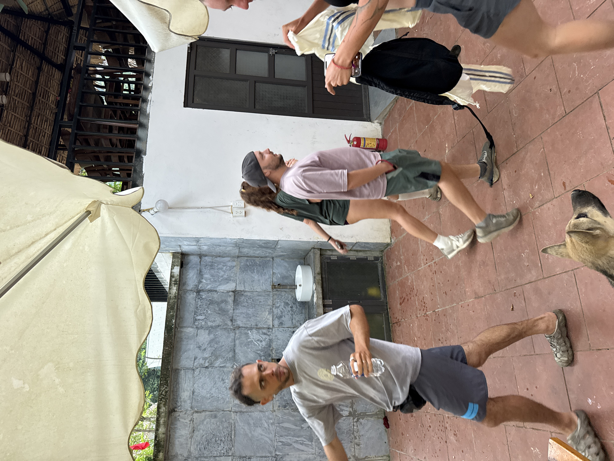 An outdoor courtyard in Cổ Lũng Commune, Vietnam, features a group of people and a dog. A man in a grey t-shirt and dark shorts holds a water bottle, walking towards the left. Behind him, a woman in a dark green t-shirt and shorts walks away from the camera. To her right, a man wearing a pink t-shirt and green shorts, with a grey baseball cap, walks forward. A woman on the far right, wearing a grey outfit, is holding a white shirt with blue stripes and a black backpack. A dog with light brown fur is visible in the lower right corner, facing left. The courtyard has a tiled floor and a stone wall. In the background, there is a building with a thatched roof and wooden railings. A fire extinguisher is visible on the wall.