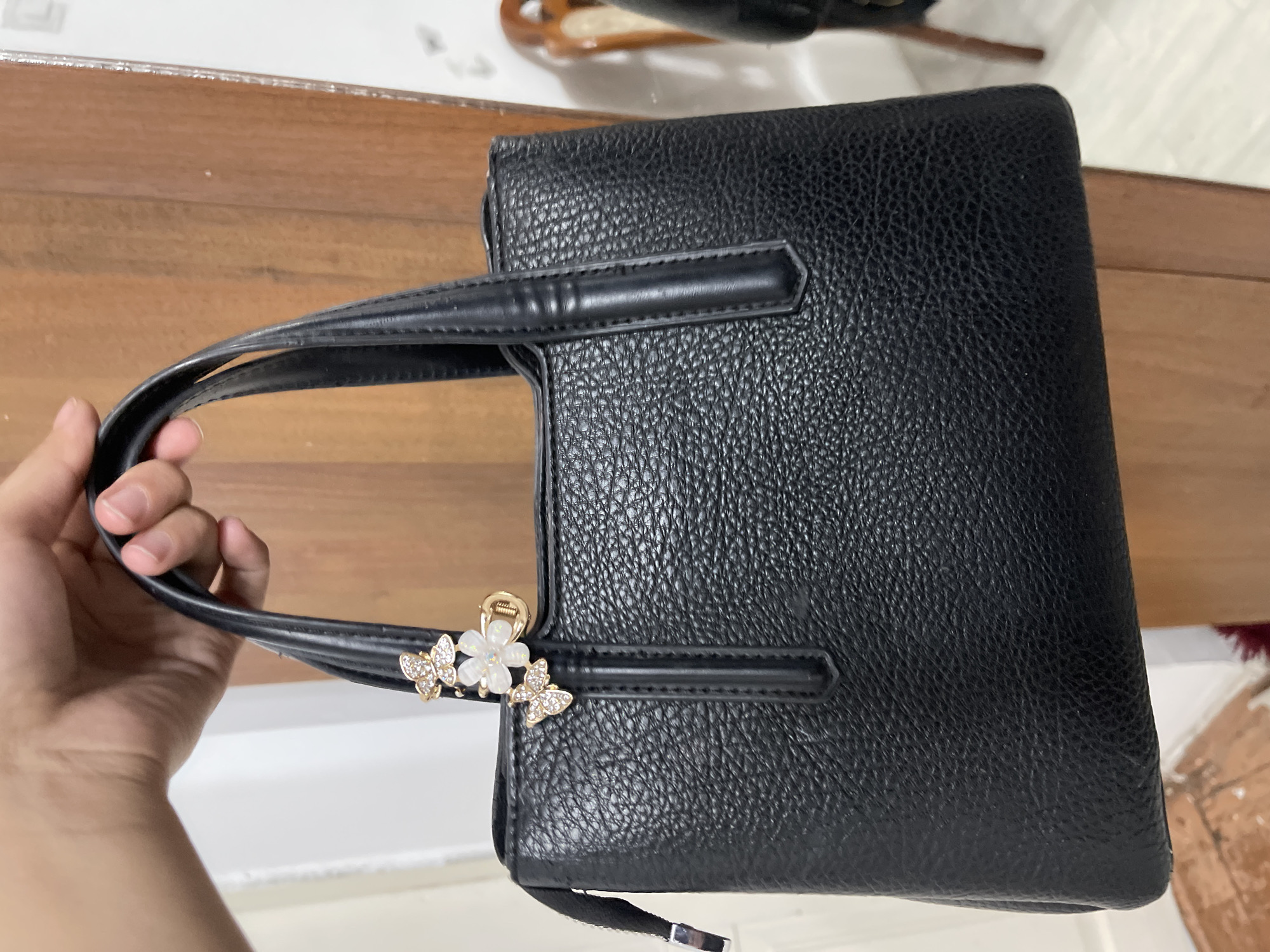 A black, textured leather handbag is held by a person's hand. The handbag has a structured, rectangular shape and two short, sturdy handles. It is adorned with a decorative charm featuring a cluster of small, sparkly butterflies and a white flower. The handbag sits on a wooden surface, likely a table, which has a light brown, grained texture. In the background, there is a white surface, possibly a wall or another table, and some out-of-focus elements suggesting an indoor environment. The lighting appears to be natural, with a soft, diffused quality, indicating it is likely daytime. The scene is static, focusing on the handbag as the primary subject. No text is visible in the image. The setting suggests a casual, domestic or retail environment in Bishkek City, Kyrgyzstan.