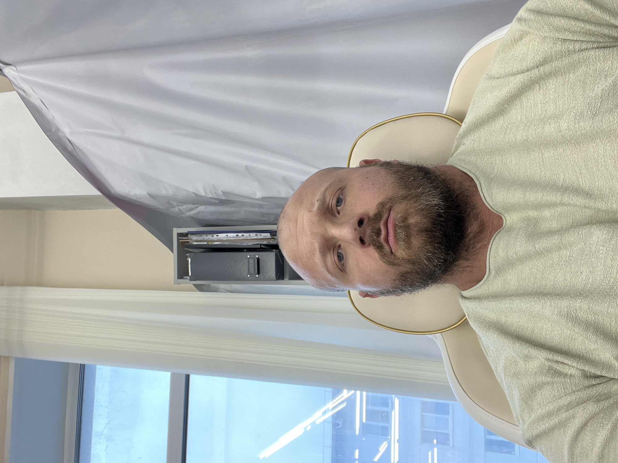 A bald, bearded Caucasian man is pictured from the chest up, seated in a plush, cream-colored chair with gold trim. He wears a light-colored, textured long-sleeved shirt. The man's gaze is directed forward, and his expression is neutral. Behind him, to the left, is a large window revealing a daytime city view with buildings and what appear to be offices or apartments. A sheer white curtain hangs to the left of the window, and a tall, light-colored cabinet or shelf unit with folders is visible to the right of the curtain, partially obscuring the man's head. To the right of the man, a large, light gray fabric screen or curtain is draped, creating a soft background. The overall setting appears to be an indoor environment, possibly an office or a waiting area. The location is specified as Chaikovsky, Russia.