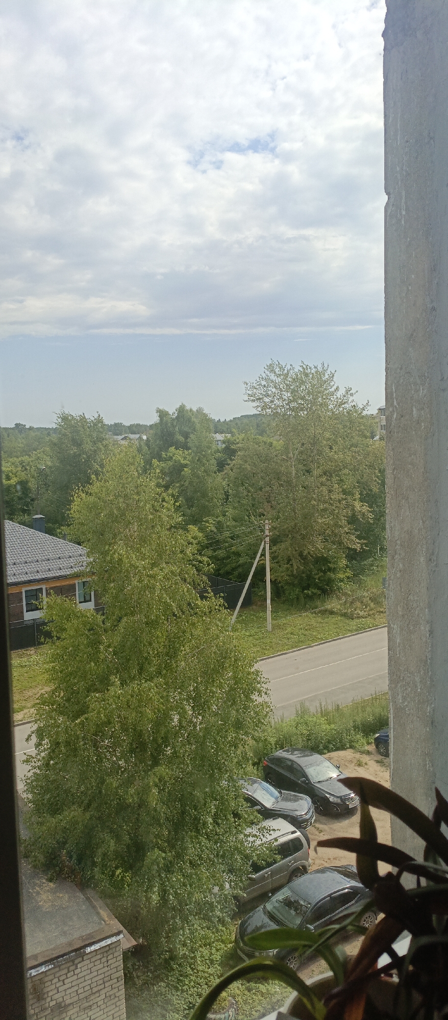 The image presents a view from a window in Sokol, Russia, overlooking a suburban landscape. In the foreground, a rough concrete pillar occupies the right side of the frame. Below the window ledge, a brick wall and part of a flat roof are visible.
The midground features a dense canopy of green trees, with a prominent birch tree with light-colored bark and a full head of leaves dominating the center-left. A paved road runs horizontally across the scene, bordered by green grass and vegetation. Several parked cars are visible along the road and on a dirt area to its right, including a dark-colored sedan in the lower right, a silver SUV, and two other dark sedans. A single utility pole with crossbars stands to the right of the birch tree, with wires extending out of frame. A modern house with a dark, pitched roof and two visible windows is situated to the left, behind a black fence.
The background shows a further expanse of trees and buildings, suggesting a residential area with some distance. The sky above is partly cloudy, with a mixture of white and gray clouds against a pale blue sky. The lighting indicates daylight, likely overcast or early morning/late afternoon given the soft light. There are no people visible in the scene.