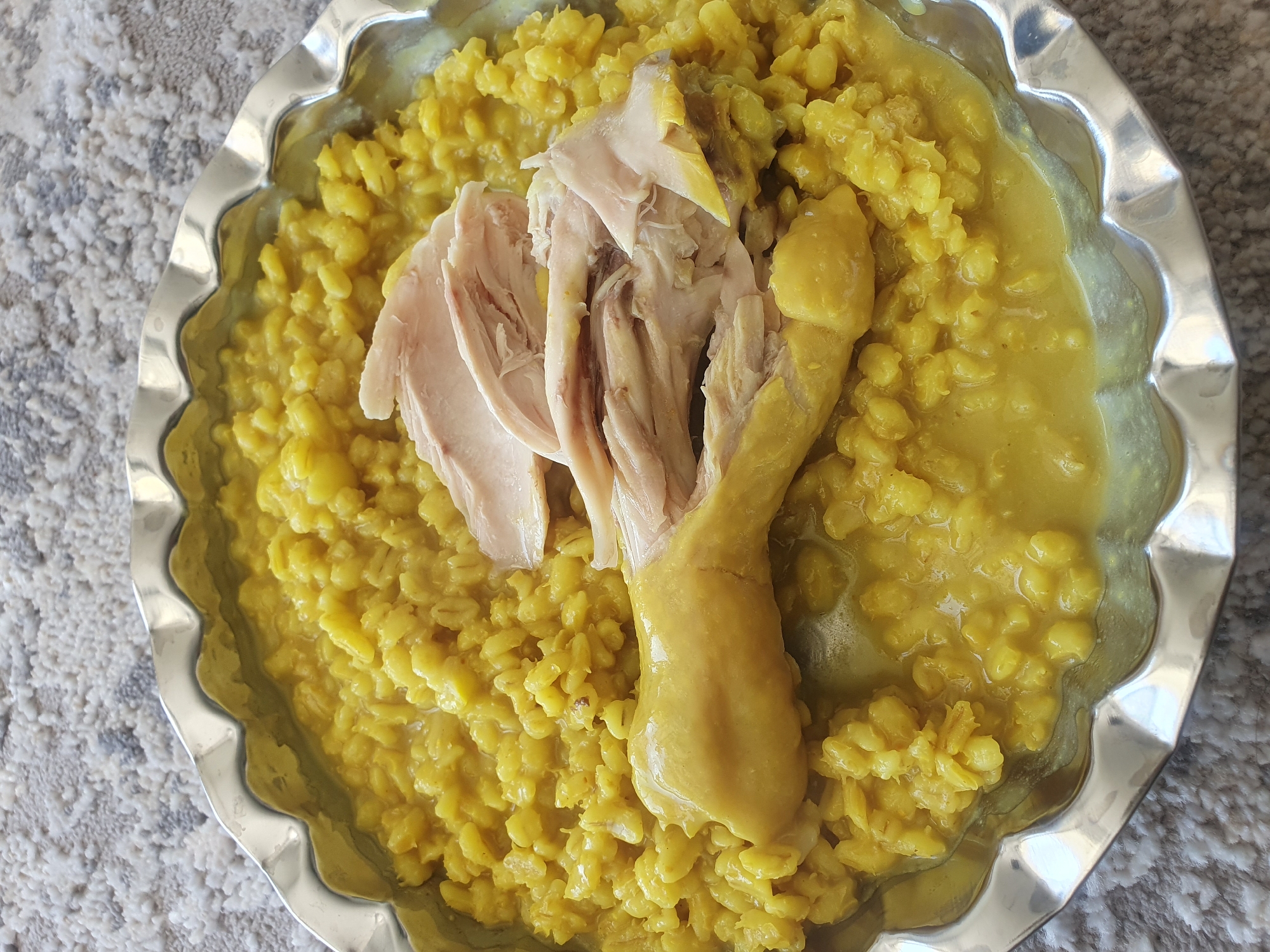 A close-up, high-angle shot shows a serving of cooked rice with shredded chicken in a decorative, scalloped silver bowl. The rice is a vibrant yellow, likely due to turmeric or saffron, and has a thick, porridge-like consistency with visible grains. A piece of cooked chicken, with some skin still attached, is placed on top of the rice, and it appears to have been shredded or pulled apart. The bowl is set on a grey and white speckled carpet. The lighting is natural, casting soft shadows. This image is of a food dish.