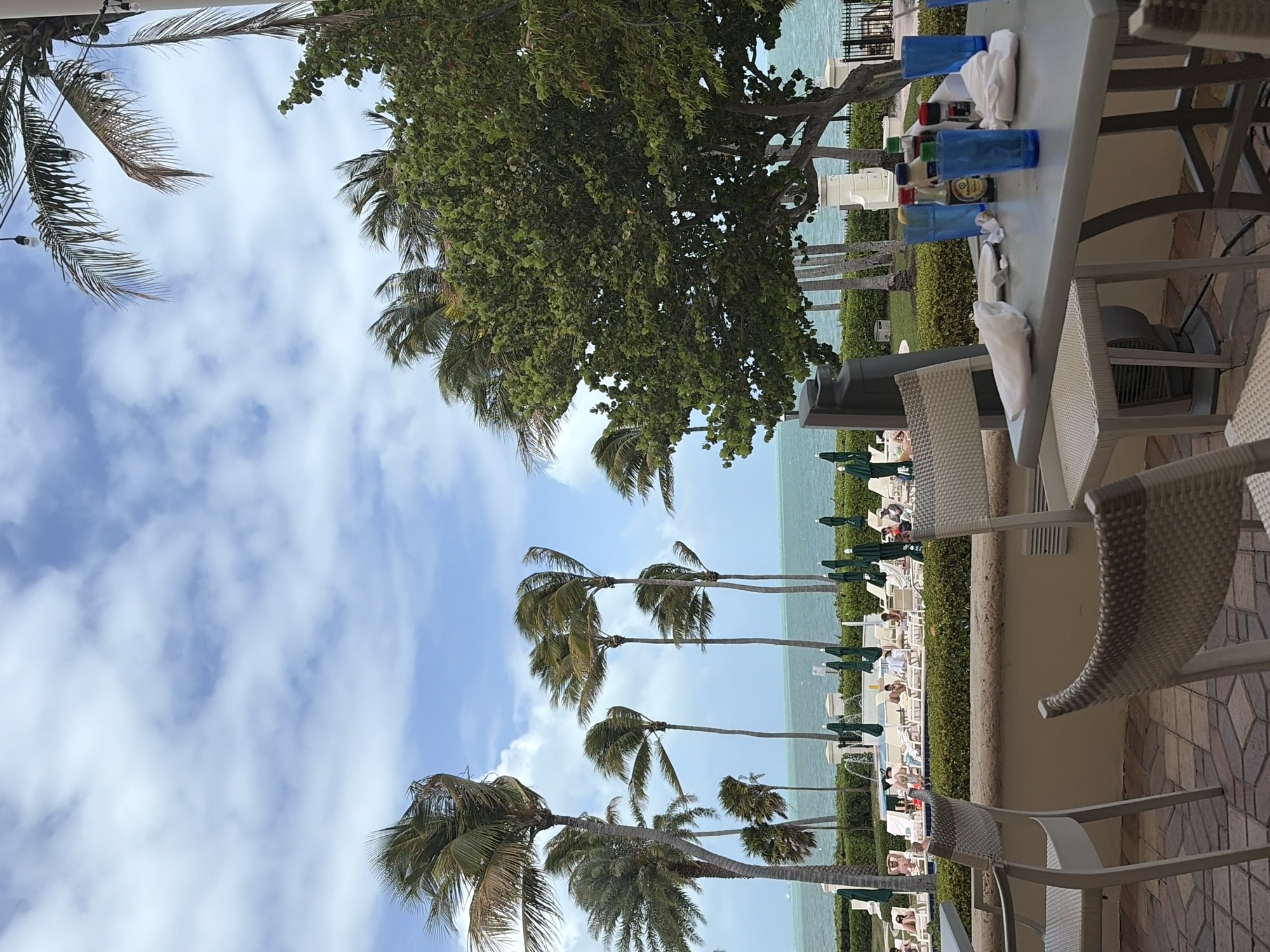 The image captures a serene outdoor scene, likely at a resort or hotel in Key Biscayne, United States of America. The foreground includes a table set with blue cups, white napkins, and various bottles, suggesting a bar or dining area. Wicker chairs are arranged around the table. Beyond the table, a landscaped area with lush greenery and palm trees leads to a view of the ocean. People are lounging on beach chairs under green umbrellas along the waterfront. The sky is partially cloudy, creating a bright and airy atmosphere. The scene depicts a relaxing and luxurious vacation setting.