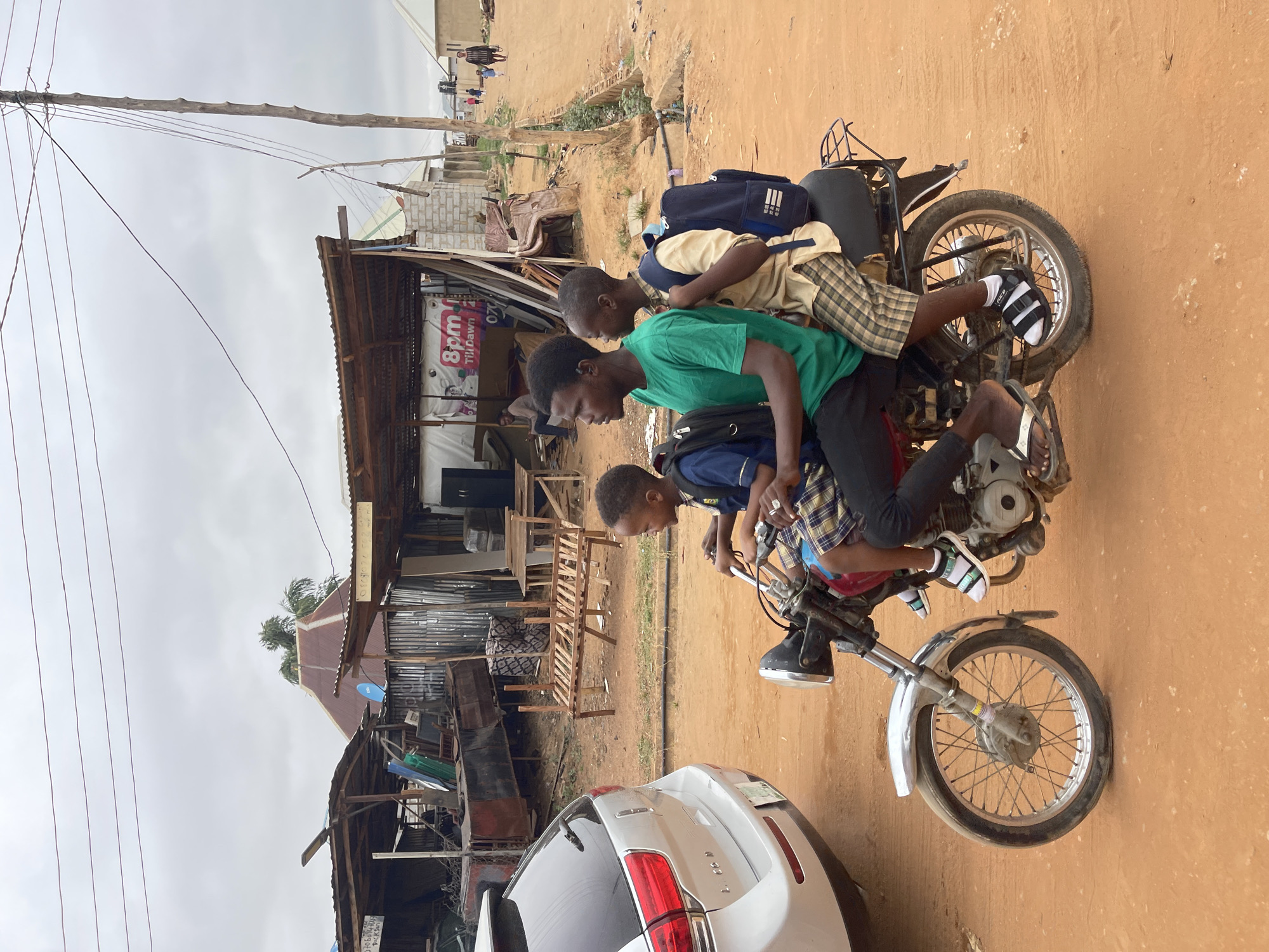 Three young men are riding on a single motorcycle in Kuchingoro, Nigeria. The motorcycle is old and the three young men are dressed in casual attire with backpacks. The location is marked by modest structures, one partially open and cluttered with furniture. A white car is parked partially within the view of the image. Overhead power lines suggest a partially developed urban area. The ground is a dusty, reddish-brown dirt.