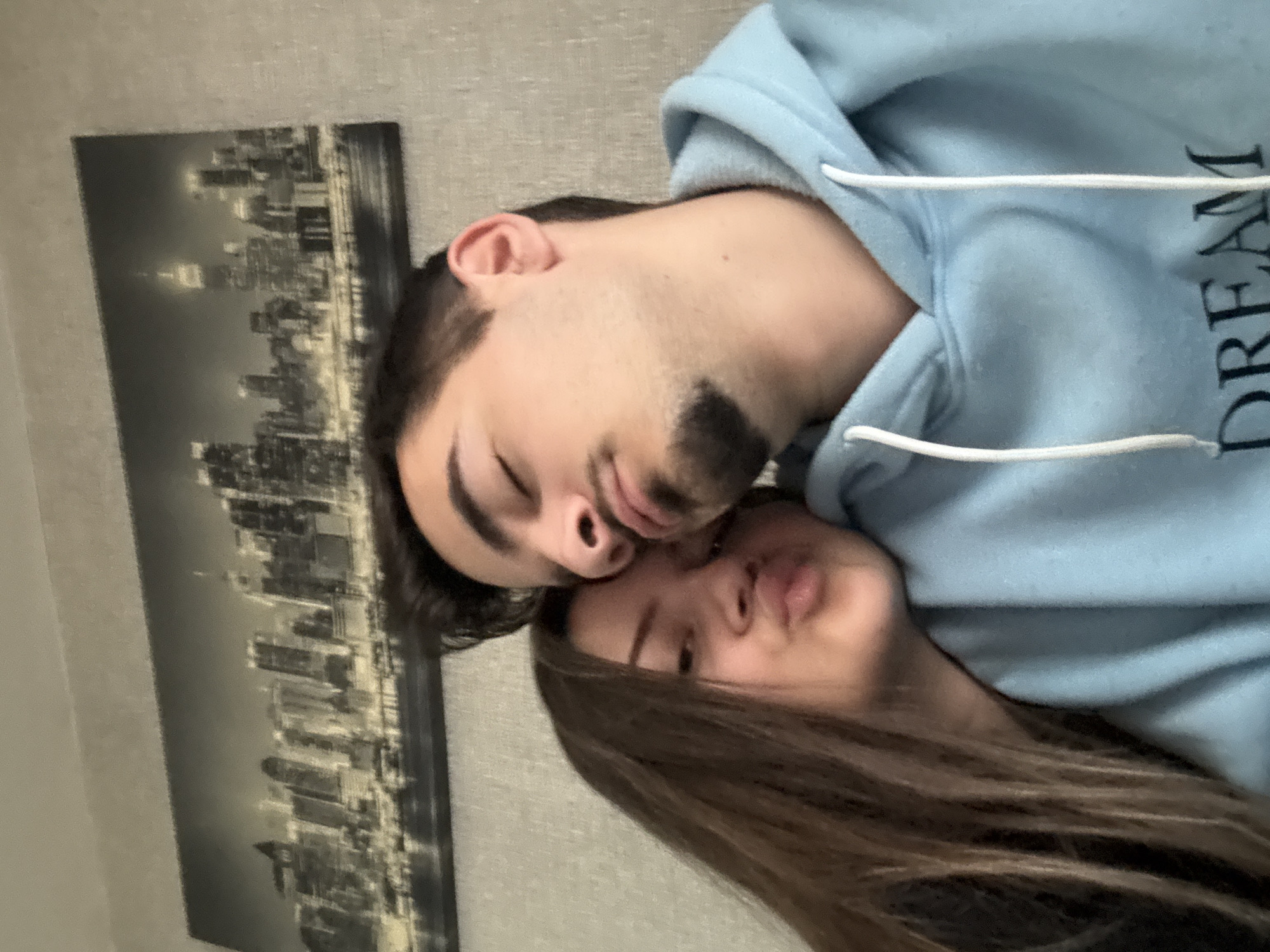 A close-up, rotated image depicts two people, a man and a woman, lying close together. The man is on top wearing a light blue hoodie with the word "DREAM" visible on the side. He has dark hair, a trimmed beard, and his eyes are closed. The woman's head is positioned underneath the man's. She has long brown hair, and her lips are pursed. Behind them, on the wall, is a black and white picture of a city skyline, resembling New York. The background wall is a neutral tone. The location is not Sofia, Bulgaria, as no elements in the picture suggest this location.