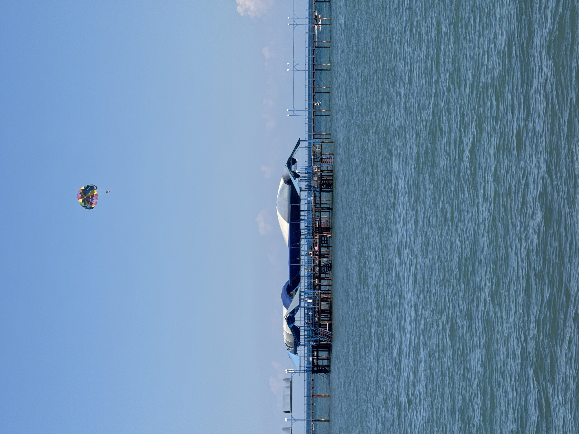 The image shows a bright, clear day at a seaside location. The sky is a vibrant, cloudless blue. In the distance, a colorful parachute with a person suspended beneath it is visible against the sky, indicating recreational activity like parasailing.
In the foreground and mid-ground, a long pier extends out over the calm, blue water. The pier has multiple covered sections with blue and white canopies, suggesting it might be a beachside establishment with seating or a place for water activities. Several people are visible on the pier, some seated and others standing, engaged in various activities. Lights are mounted on poles along the pier.
The water is a deep blue with gentle ripples, reflecting the clear sky. To the right of the pier, a small boat or kayak can be seen on the water. The overall scene depicts a sunny day at a resort or beach area, likely in a coastal city, where tourists can enjoy water sports and relax by the sea. The location is identified as Sary-Oy, Kyrgyzstan, which is situated on the northern shore of Issyk-Kul Lake.