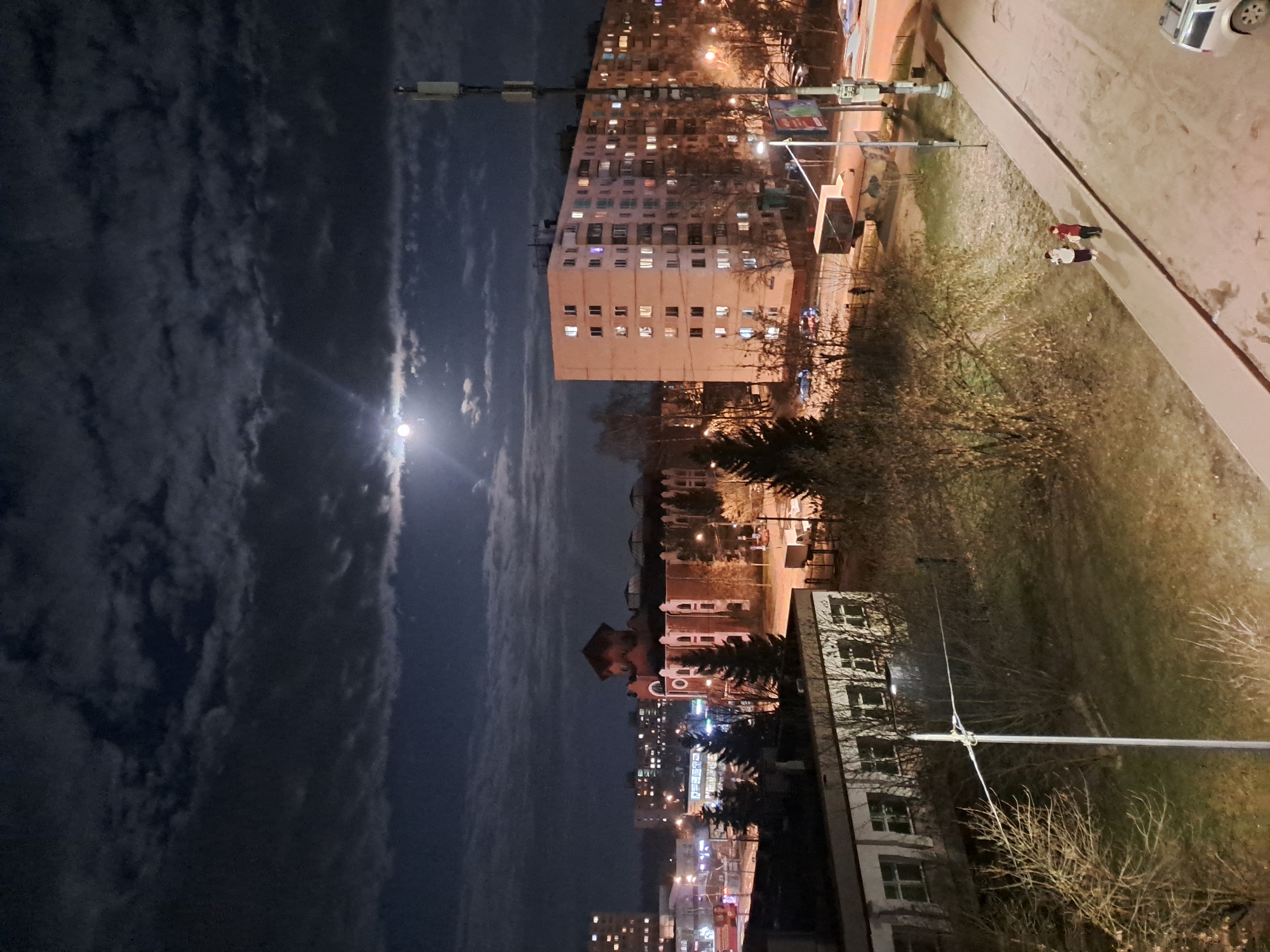The image shows an urban night scene in Elektrostal, Russia. A bright full moon is visible in a sky partially obscured by clouds. Below, a tall, rectangular apartment building with numerous illuminated windows dominates the midground. Further in the background, other buildings are visible with streetlights creating a network of illumination. A sidewalk runs along the right side of the image, where two individuals are walking. A parked car is visible in the top right corner. Trees and vegetation are scattered throughout the scene.