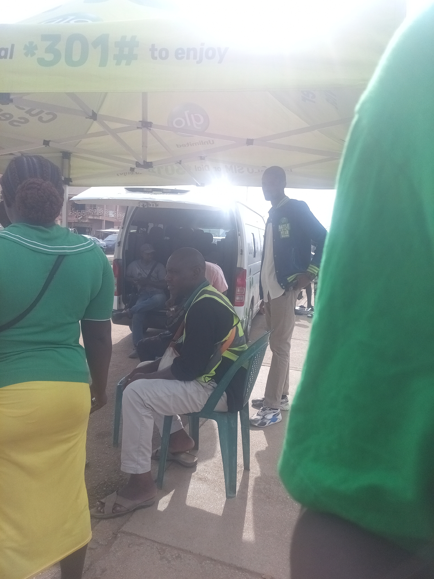 The image captures an outdoor scene, likely in Jos, Nigeria, under a branded tent. The tent's branding includes the text "al *301# to enjoy". Several individuals are present, with varying degrees of visibility. In the foreground, a person wearing a bright green top and a yellow skirt is partially visible from the back, facing away from the camera. To the left, another individual wearing a dark jacket over a light-colored shirt and khaki pants is standing, with their hands in their pockets. In the middle ground, a white van is parked, with its rear doors open. Inside and around the van, several people are seated or standing. One man in the mid-ground, wearing a black jacket with a reflective yellow vest over it, is seated on a green plastic chair. He is looking to his right. Another person in a gray t-shirt and blue jeans is visible seated inside the van. The ground appears to be a mix of pavement and gravel. The lighting suggests it is daytime, with sunlight creating bright highlights and shadows. The overall atmosphere seems casual, possibly related to an event or service being offered under the tent.