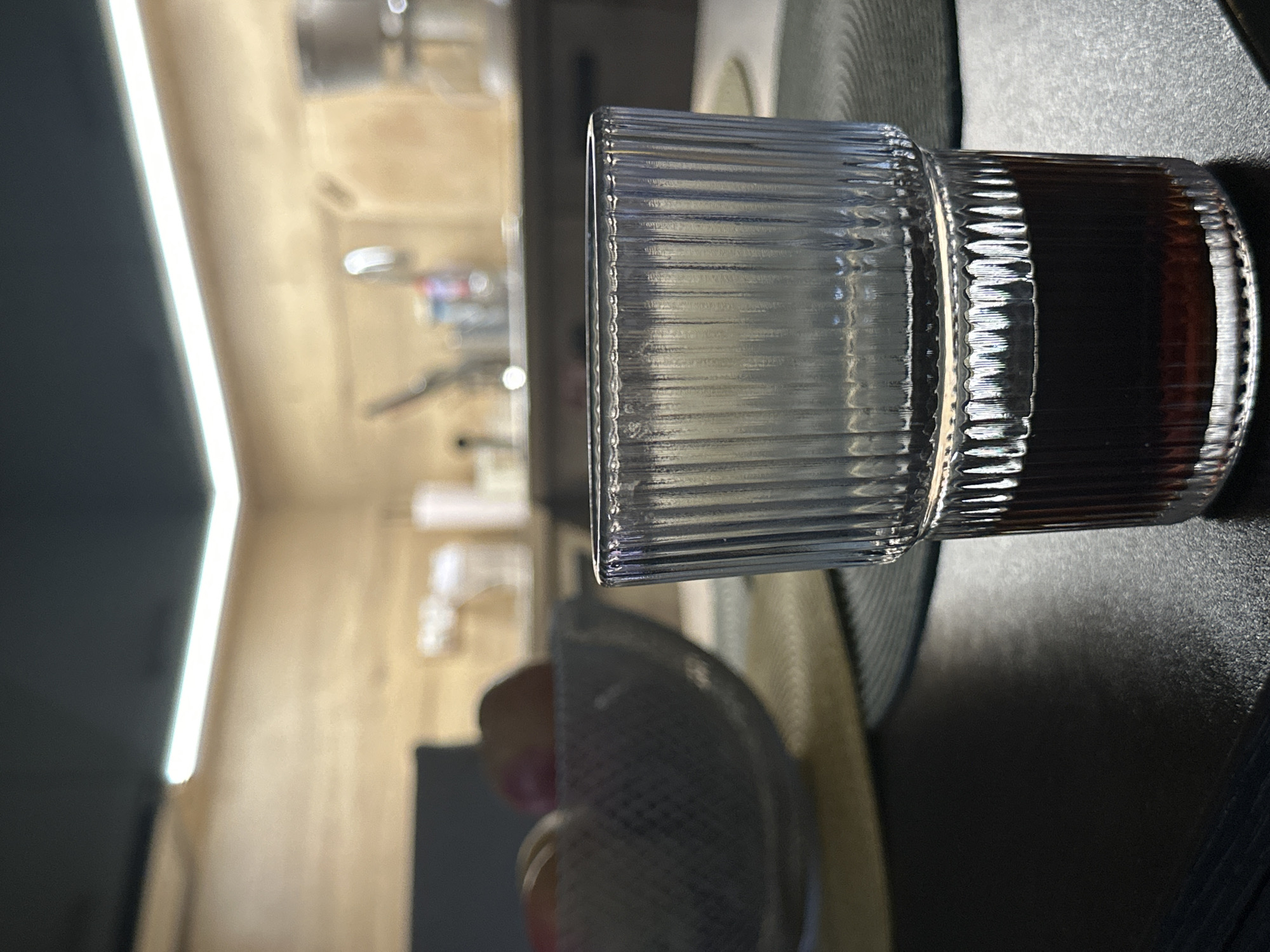 The scene depicts an indoor environment, specifically a kitchen or dining area in Sofia, Bulgaria, viewed from a low angle. In the immediate foreground, a clear, vertically ribbed glass tumbler, approximately half-filled with a dark brown or black liquid, rests on a dark, textured tabletop. To the left and slightly behind the glass, a transparent or translucent bowl containing several round, reddish-brown fruits (possibly apples) is situated on a light-colored, circular placemat, which itself lies on a darker, woven placemat. The background is significantly out of focus, revealing a modern kitchen space. This area features light-colored wooden wall panels or cabinetry illuminated by a bright, linear LED strip light positioned at the intersection of the wall and a dark, possibly black, ceiling. Vaguely discernible kitchen fixtures include a metallic sink faucet, various containers on a countertop, and a white coffee maker towards the far right. No individuals are present, and no specific actions or interactions are depicted.