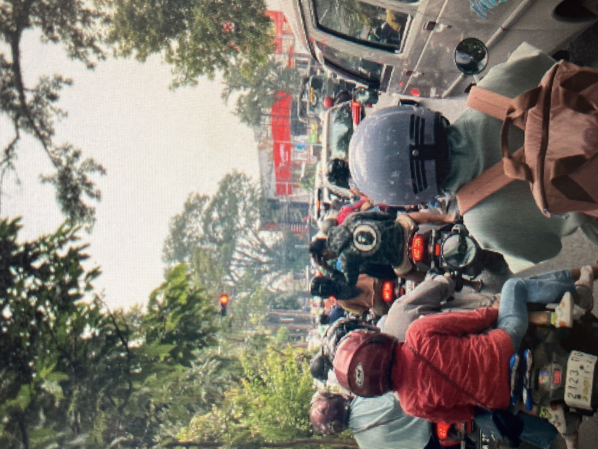 The image depicts a traffic jam in the city of Sejahtera, Indonesia. Numerous motorcycles and some cars are stopped on a road lined with trees and buildings. The perspective is from behind several people on motorcycles. One person in the foreground is wearing a red shirt, blue jeans, and a red helmet, sitting on a motorcycle with a license plate visible. To their right, another person wears a light green shirt and a grey helmet, carrying a tan backpack. In the background, more motorcycles and vehicles are visible, with buildings and bright red signage in the distance. A traffic light is illuminated red overhead. The overall atmosphere suggests a busy urban environment during daytime.